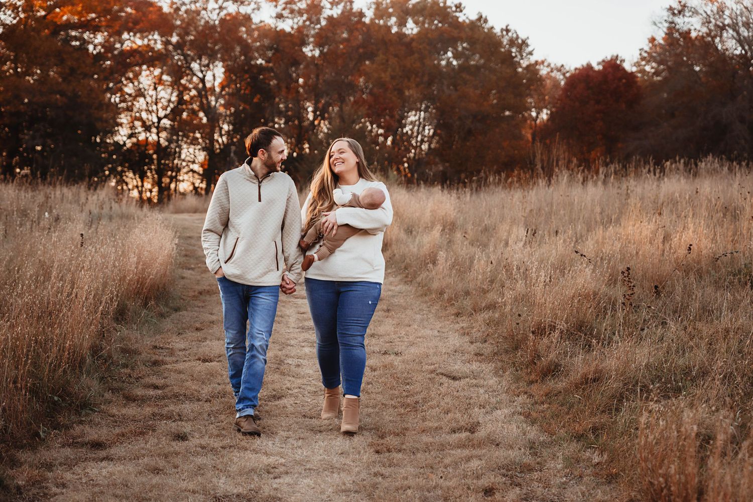 Happy couple walks hand-in-hand through autumn field, mother cradling newborn baby.