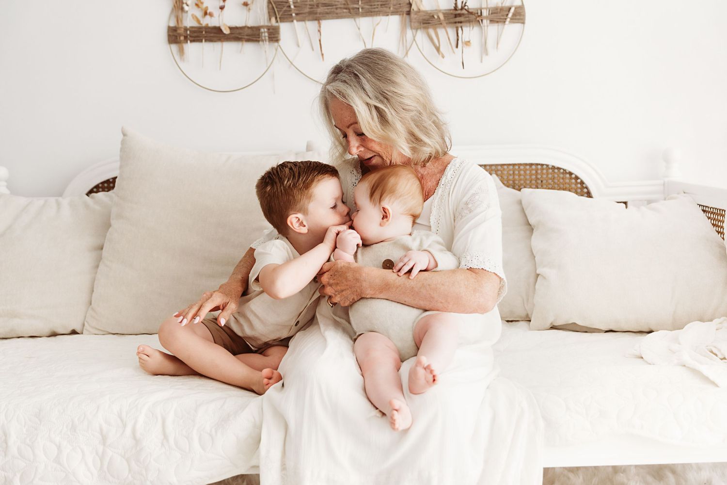 A grandmother sits on a white sofa lovingly holding her two young grandchildren in a bright room.