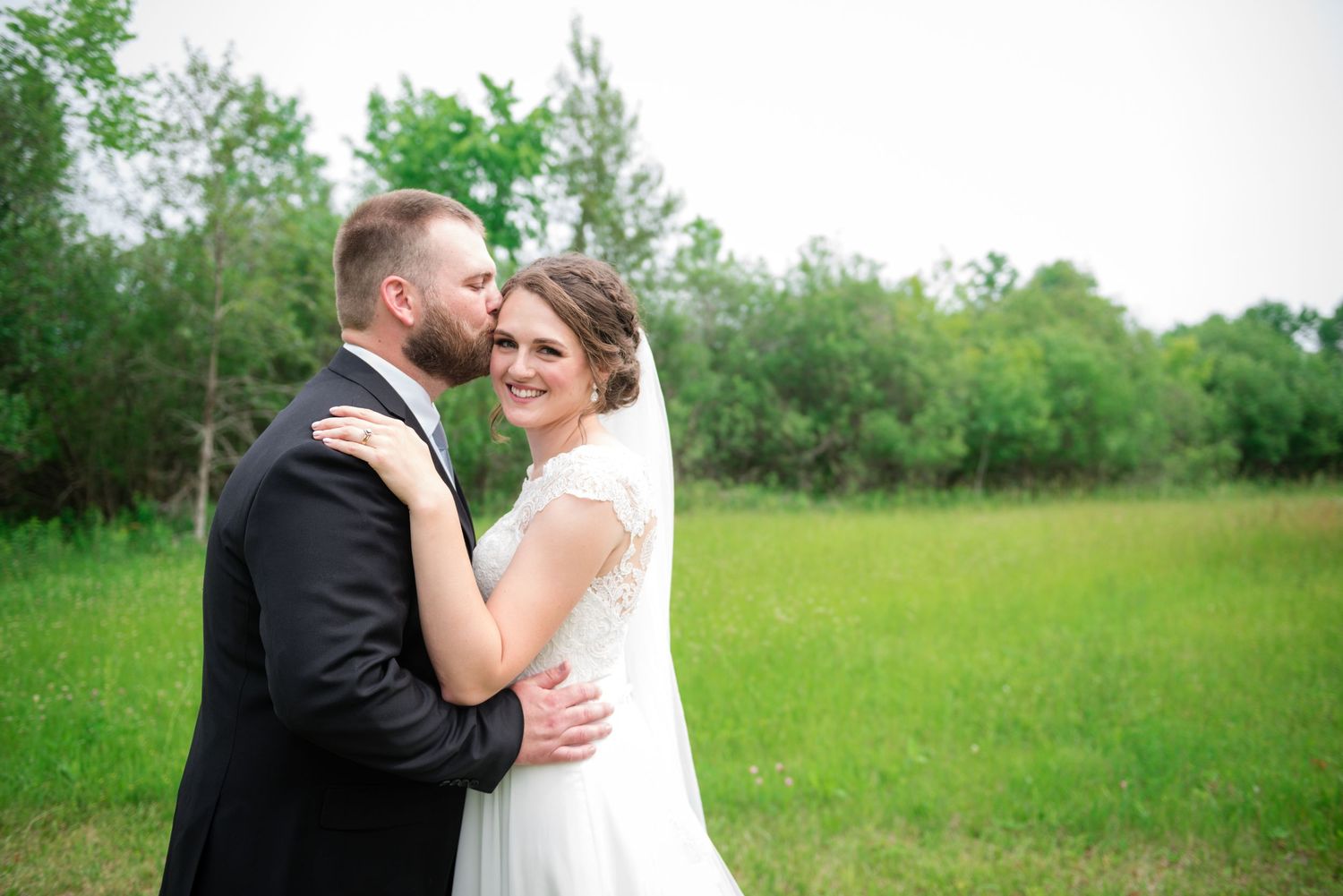 Newlywed couple embracing in a lush green field, groom kissing bride's cheek as she smiles.