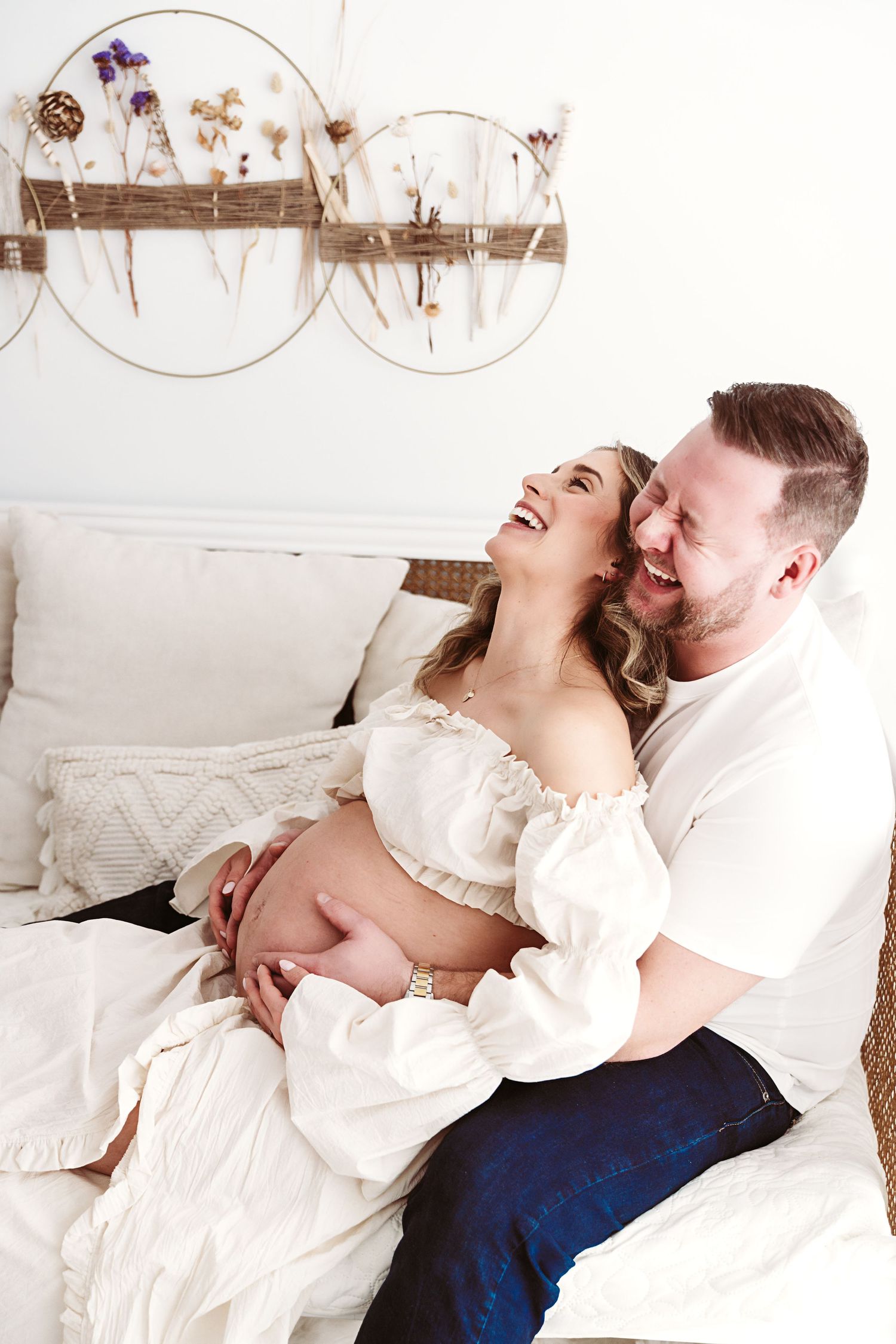 Happy pregnant couple laughing together on a white couch in a boho-decorated room.