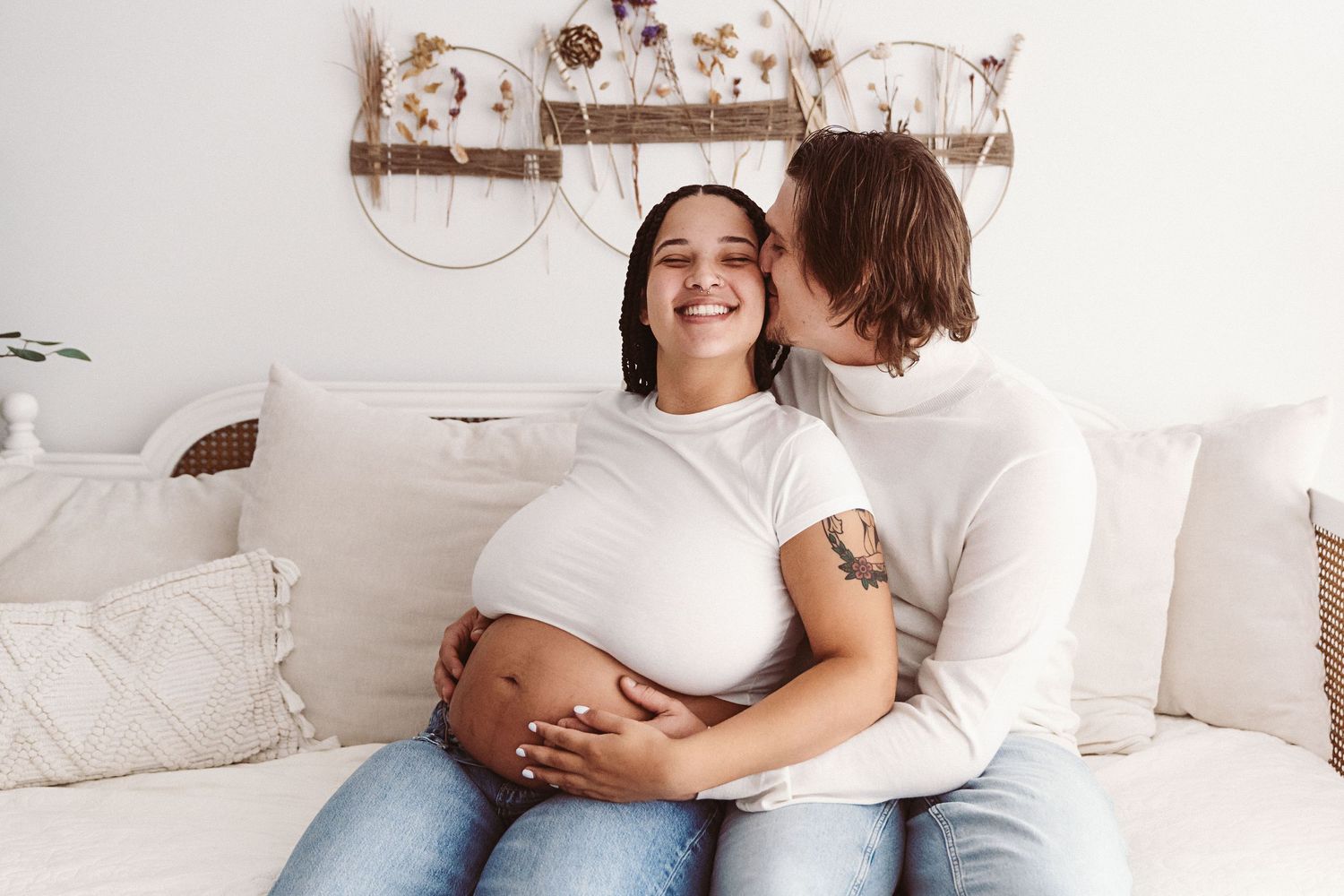 Happy pregnant woman smiling as partner kisses her cheek while cradling bump on a cozy white sofa.