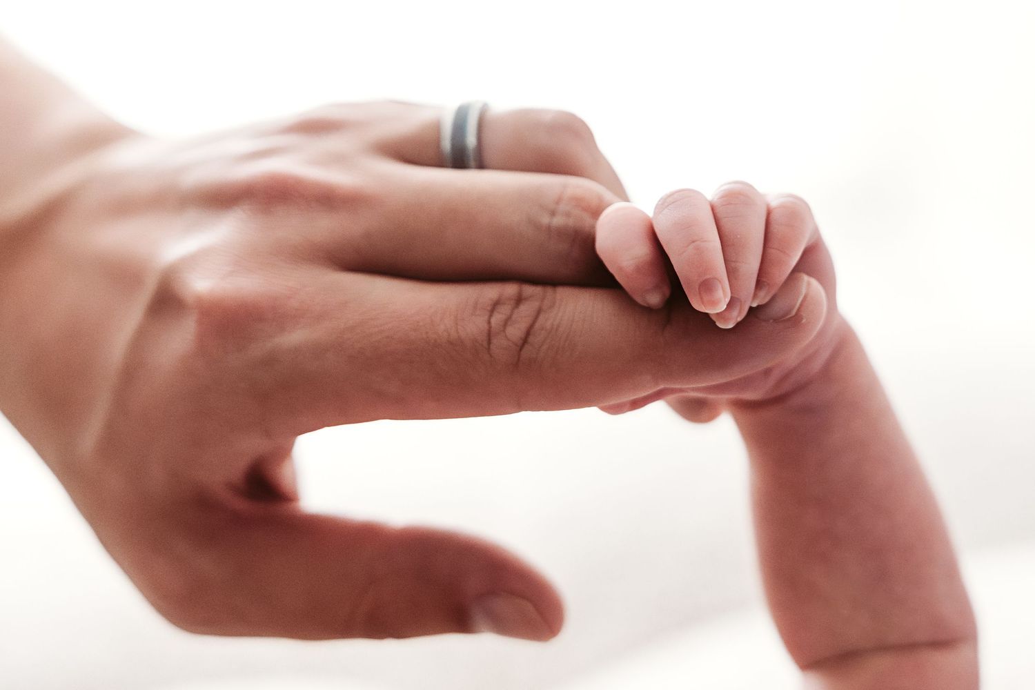 A tiny newborn baby's hand grasps an adult's finger wearing a silver wedding band.