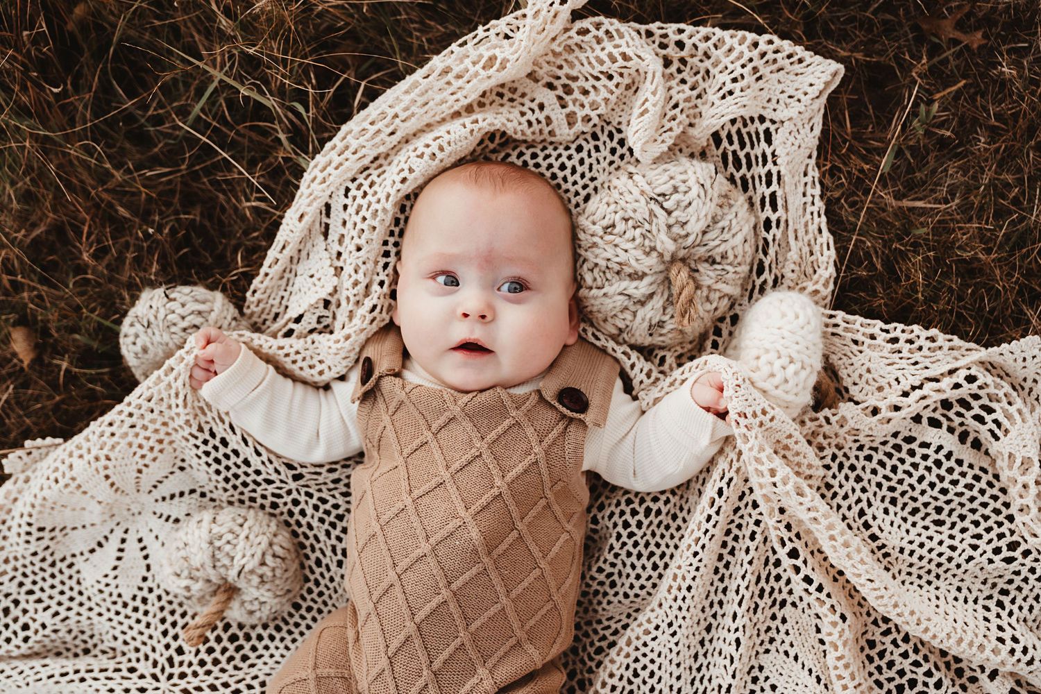 Adorable baby in brown knit romper lies on cream crochet blanket outdoors.