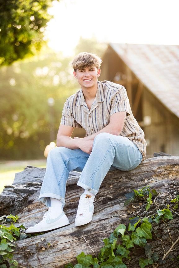 Smiling young man in striped shirt and jeans sitting on a log outdoors in golden sunlight.