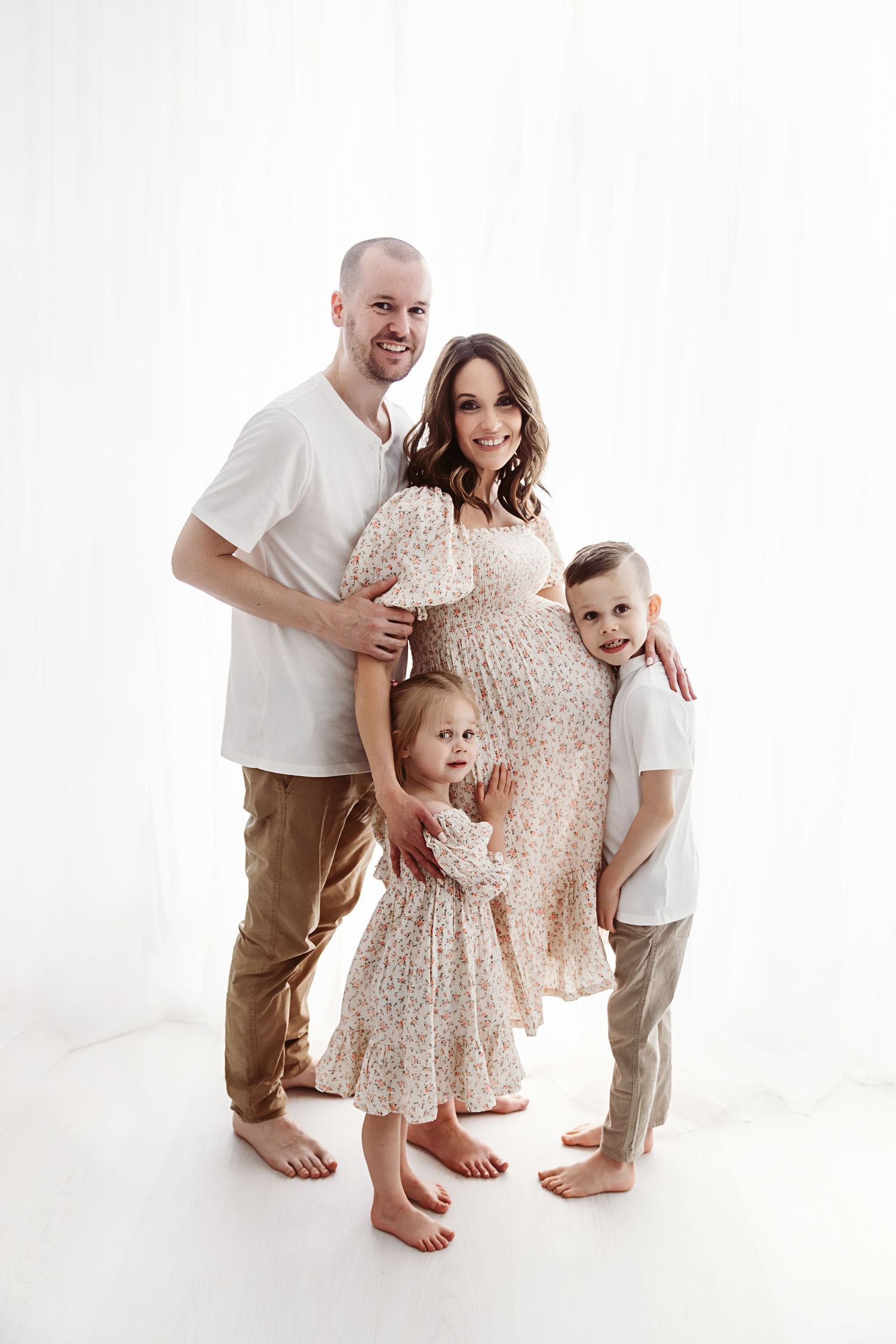 Happy family of four with pregnant mother posing together in a bright white studio setting.