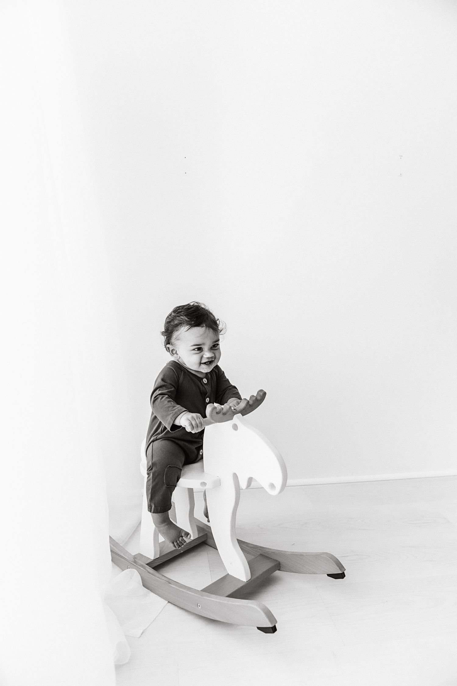 Black and white photo of baby riding a wooden rocking moose in bright white room.