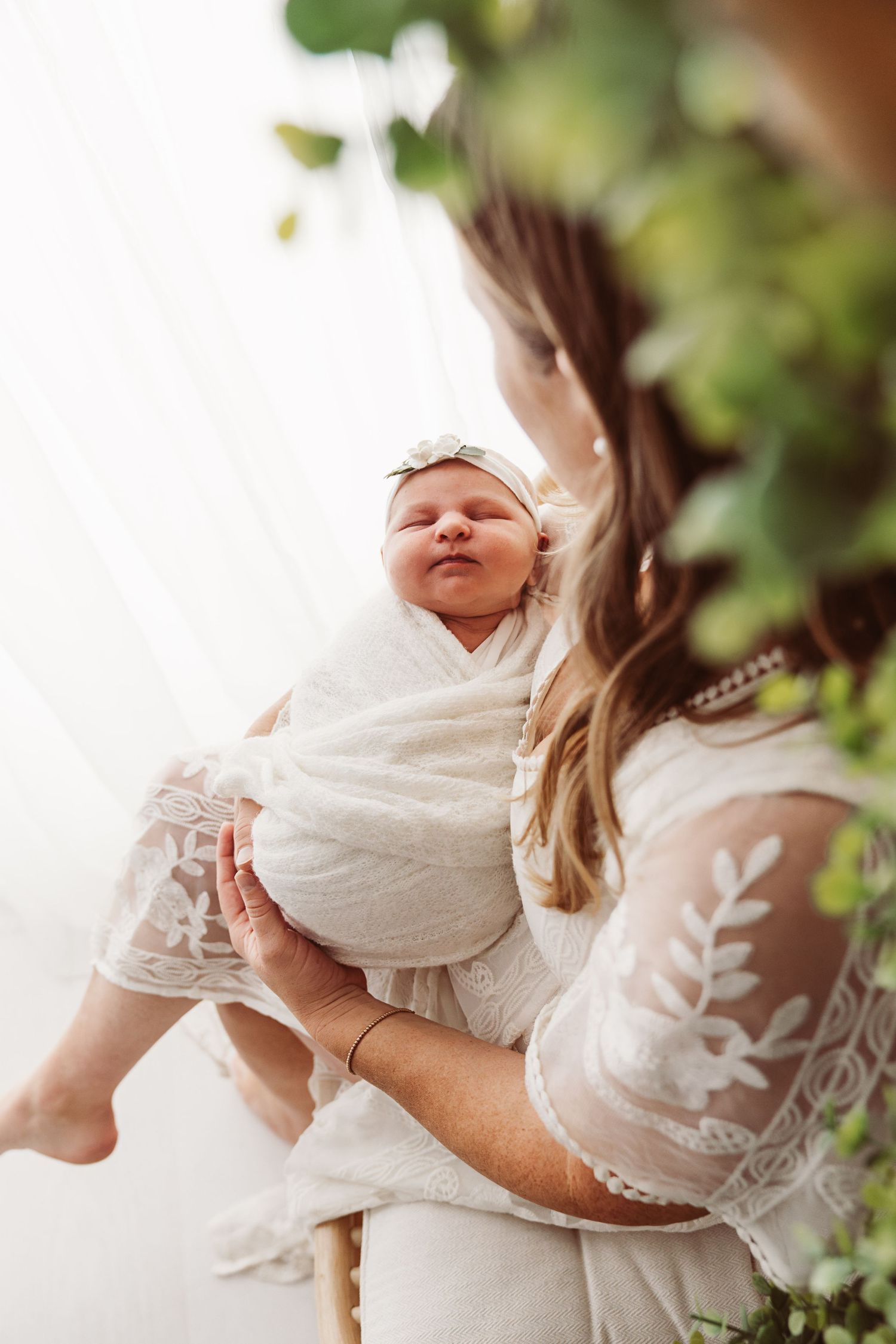 Mother in white lace dress holding her swaddled newborn baby girl surrounded by green foliage.