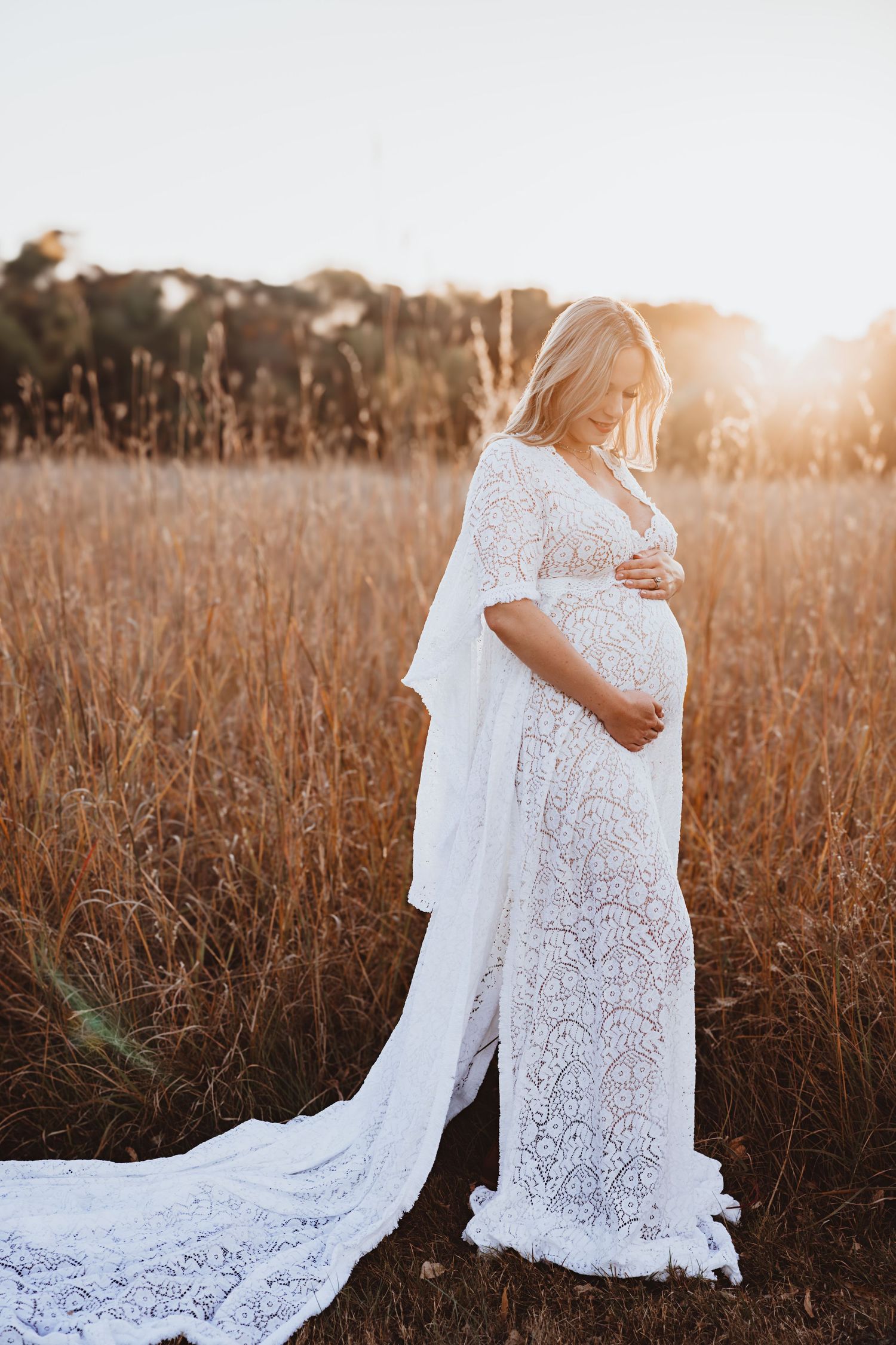 Pregnant woman in white lace maternity gown standing in golden field at sunset.