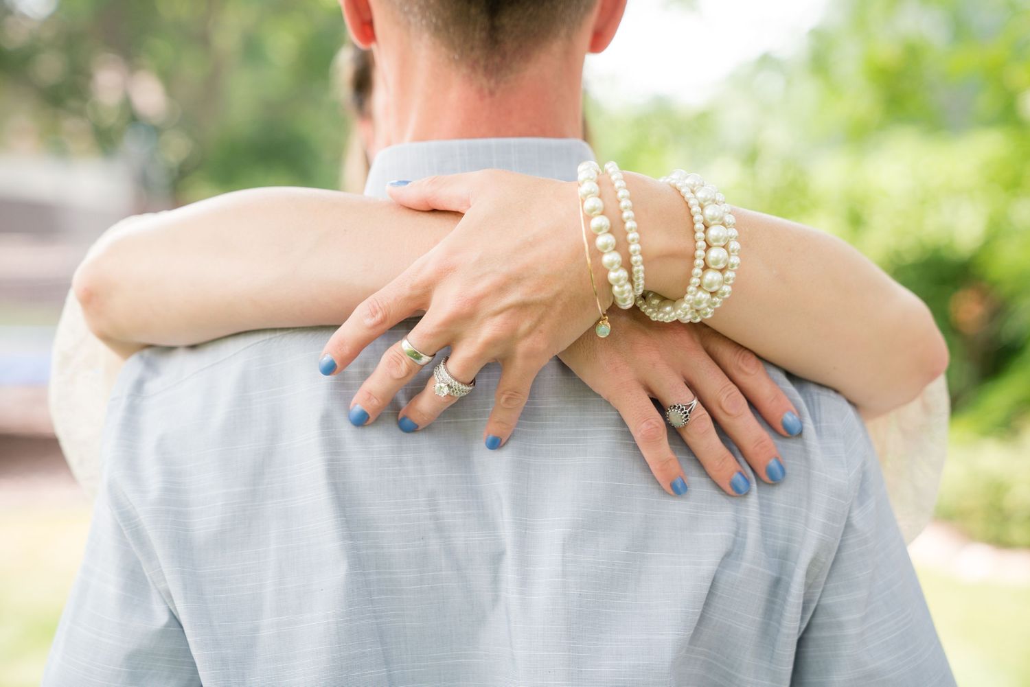 A woman with blue nails and pearl bracelets wraps her arms around a man's shoulders outdoors.