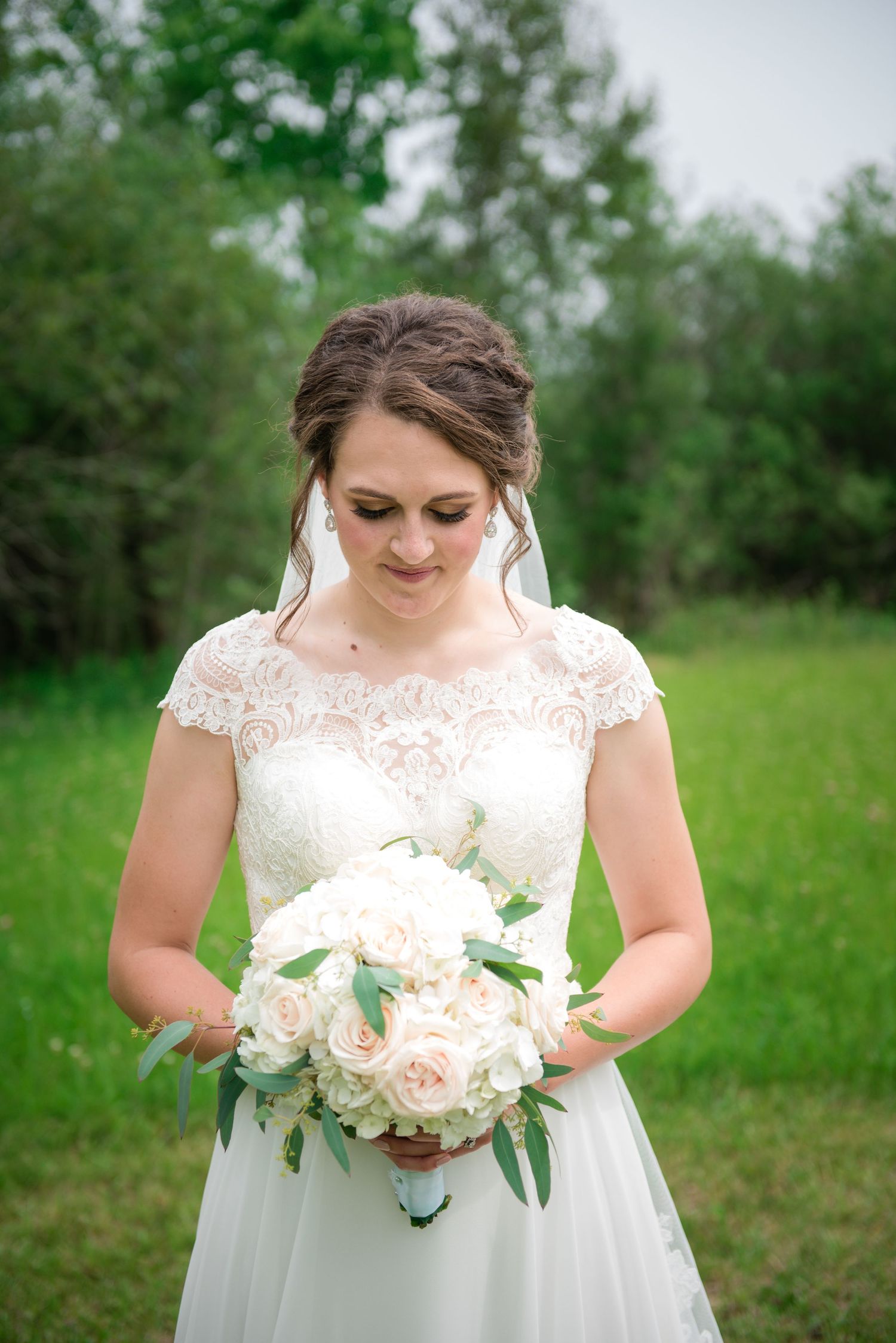 Bride in lace cap sleeve wedding dress gazing at white rose bouquet in lush green outdoor setting.