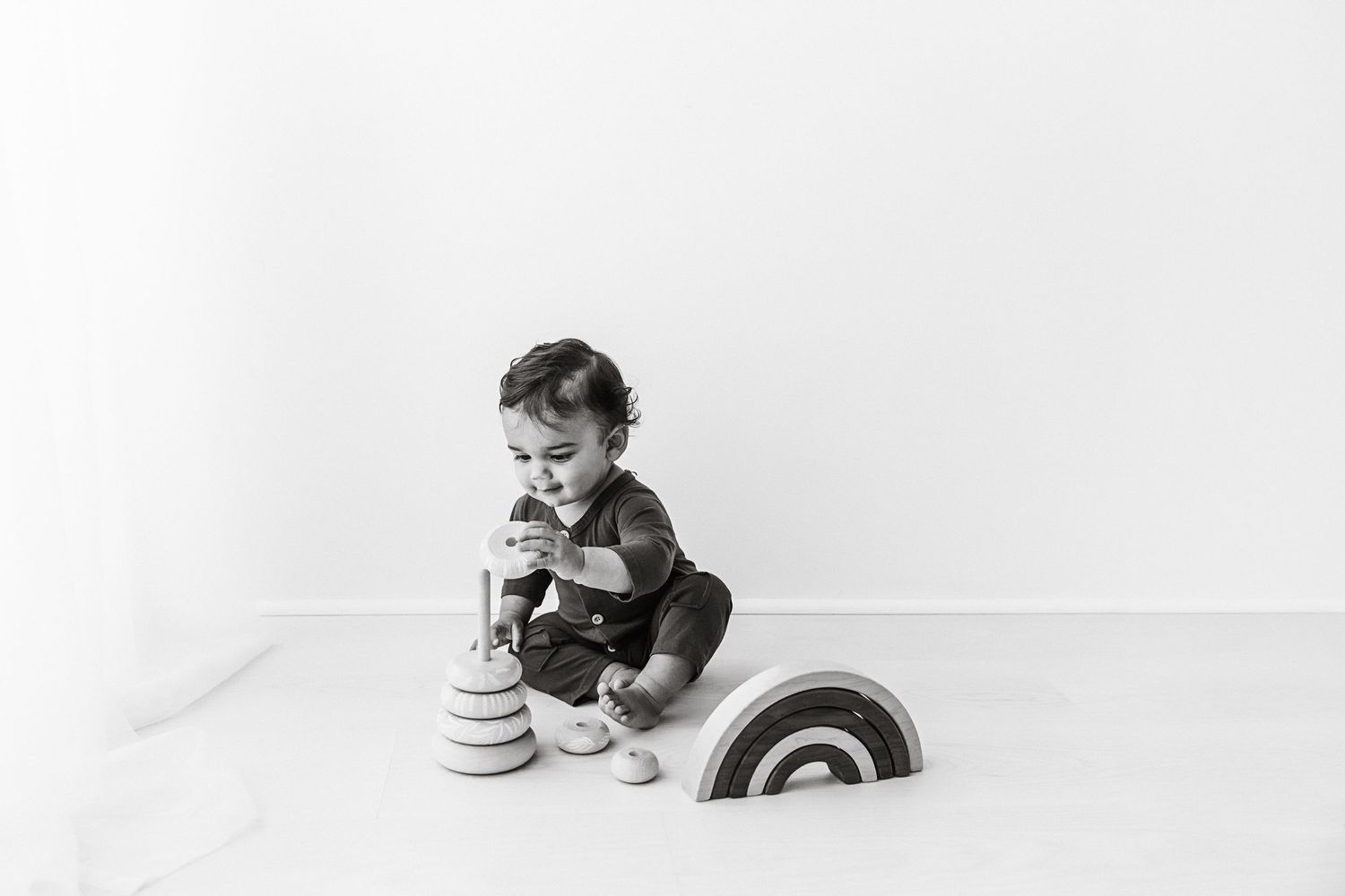 Black and white photo of baby playing with stacking ring toys on white floor.
