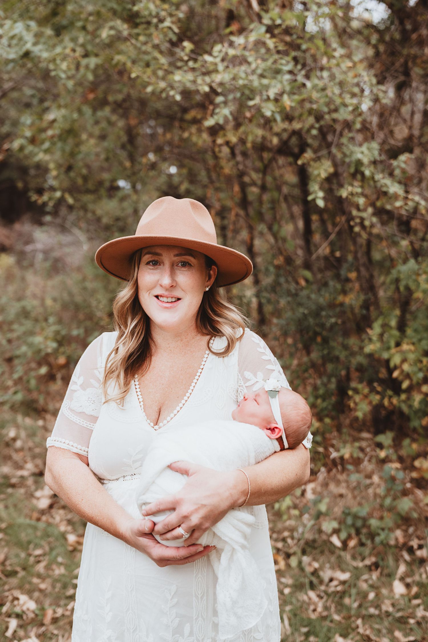 Smiling mother in white lace dress and tan wide-brim hat holding newborn baby outdoors in autumn forest.