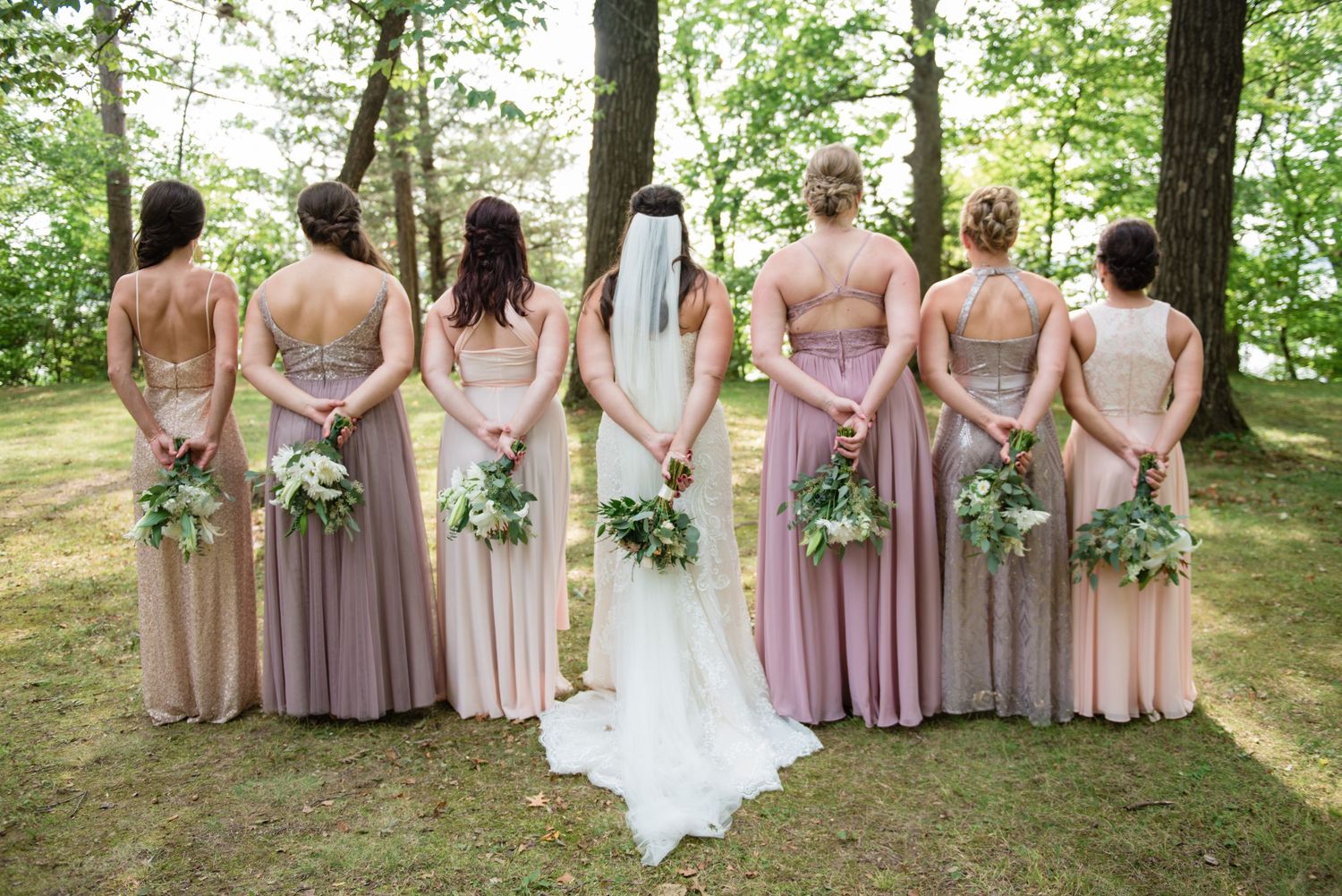Bride and six bridesmaids in mismatched mauve dresses standing with bouquets facing trees.