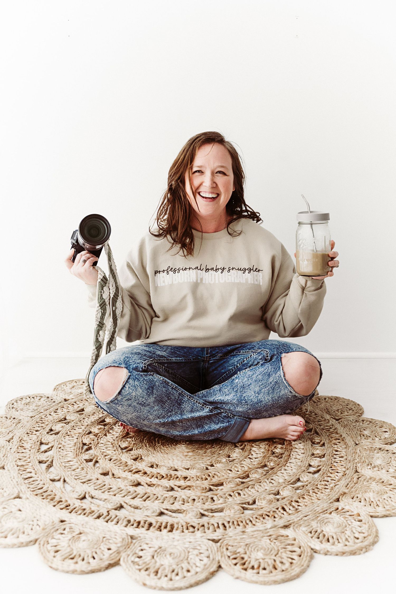Laughing woman in professional baby snuggler sweatshirt sitting on woven rug, holding camera and mason jar drink.