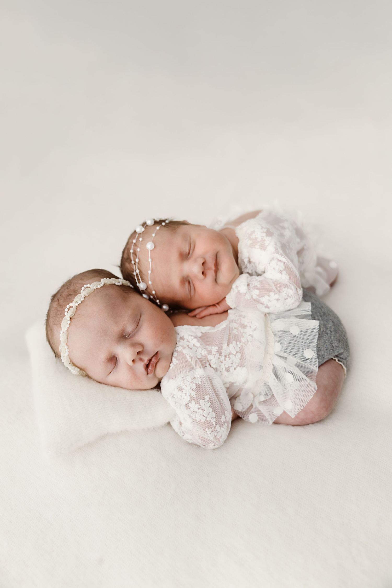 Two sleeping newborn twins cuddled together wearing delicate white lace outfits and headbands.