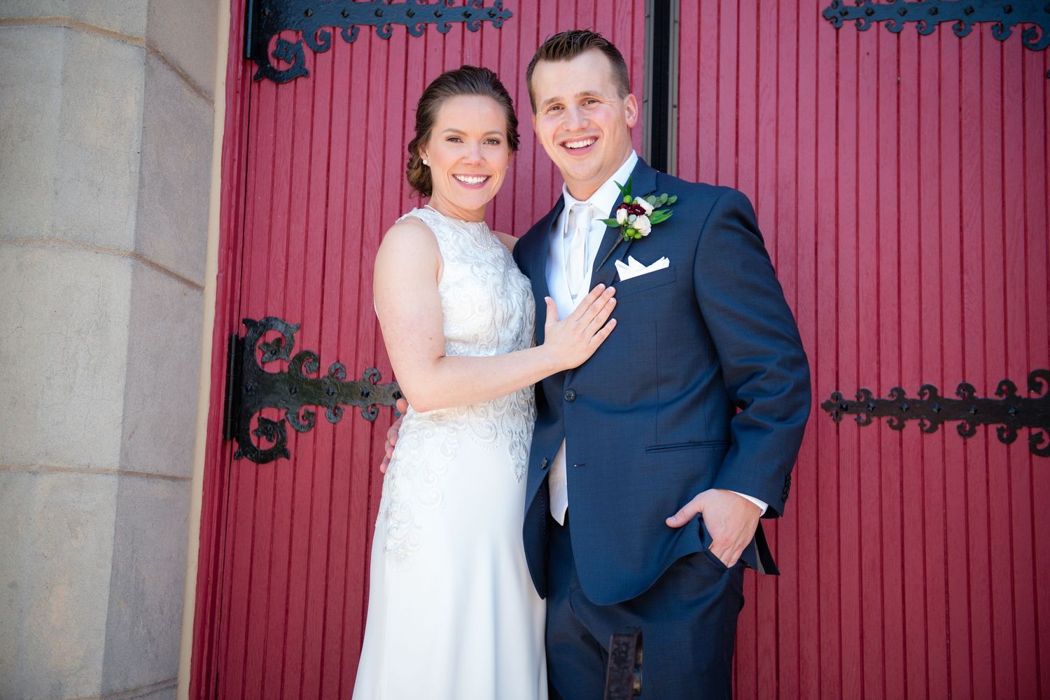Smiling bride and groom posing together in front of elegant red doors, groom wearing navy suit.