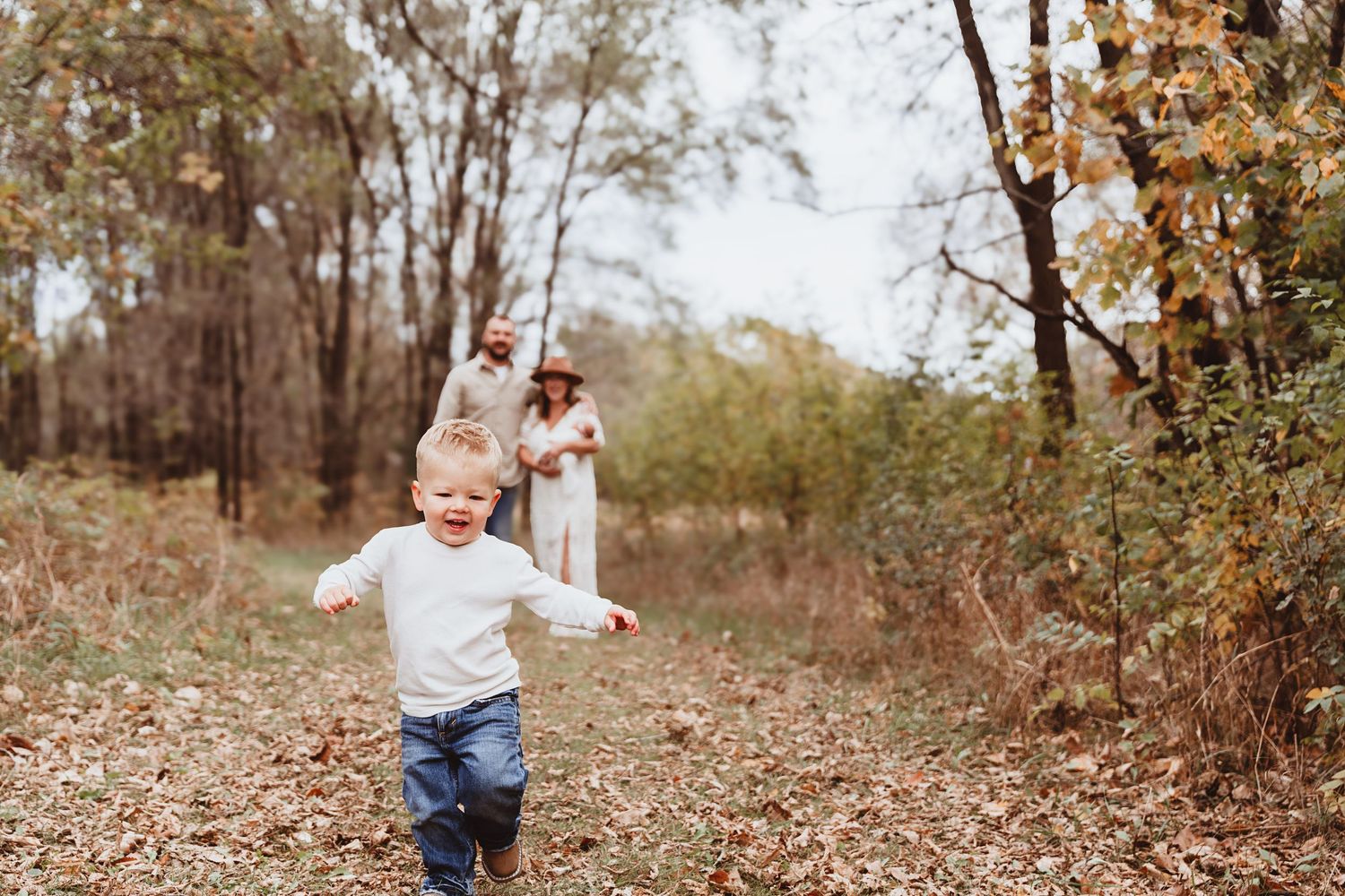 Happy toddler running with arms outstretched on autumn forest path while parents watch in background.