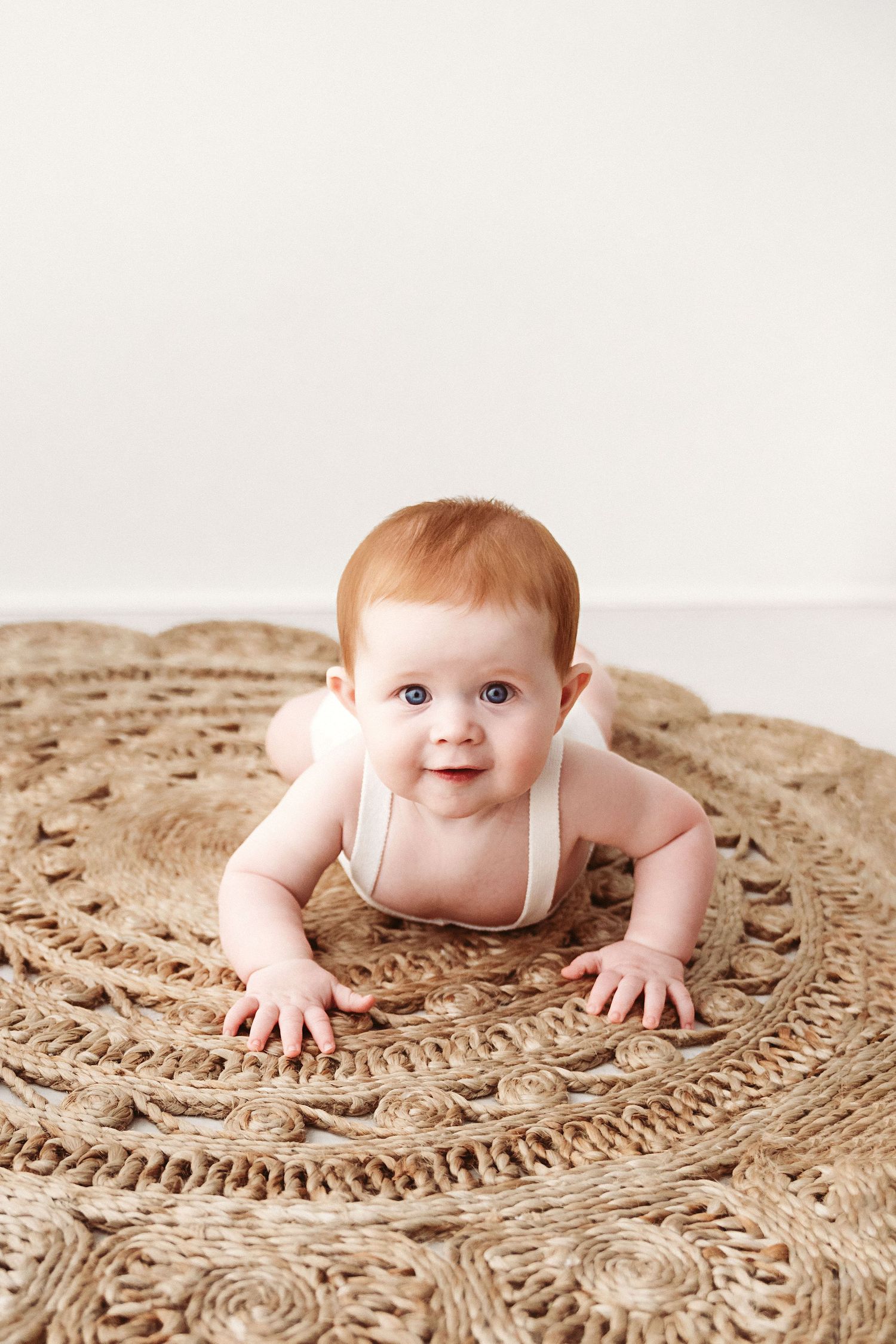 Smiling red-haired baby crawling on a round woven jute rug against a white background.