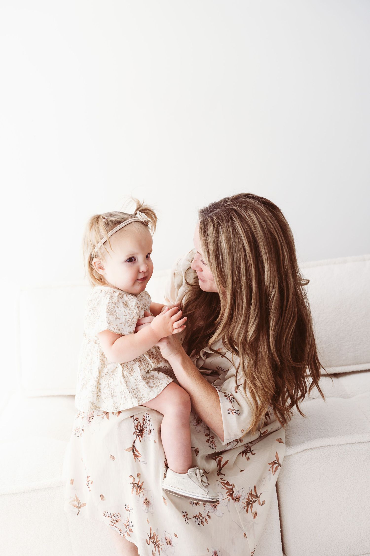 Mother holding baby girl in matching floral dresses, sharing a tender moment on a white couch.
