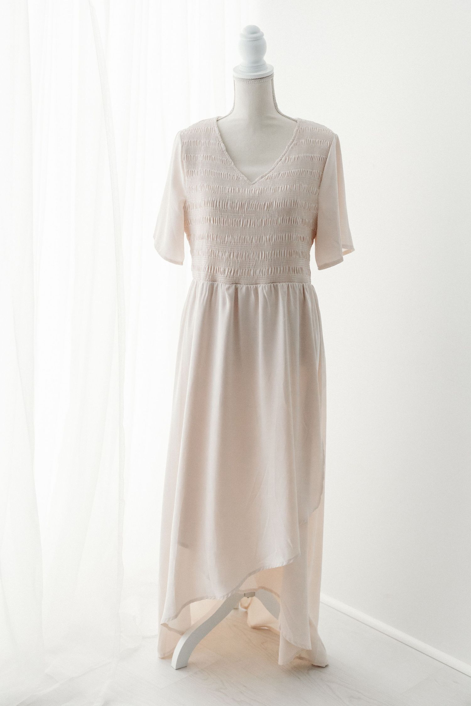 Blush pink V-neck maxi dress with smocked bodice and asymmetrical hem on mannequin.