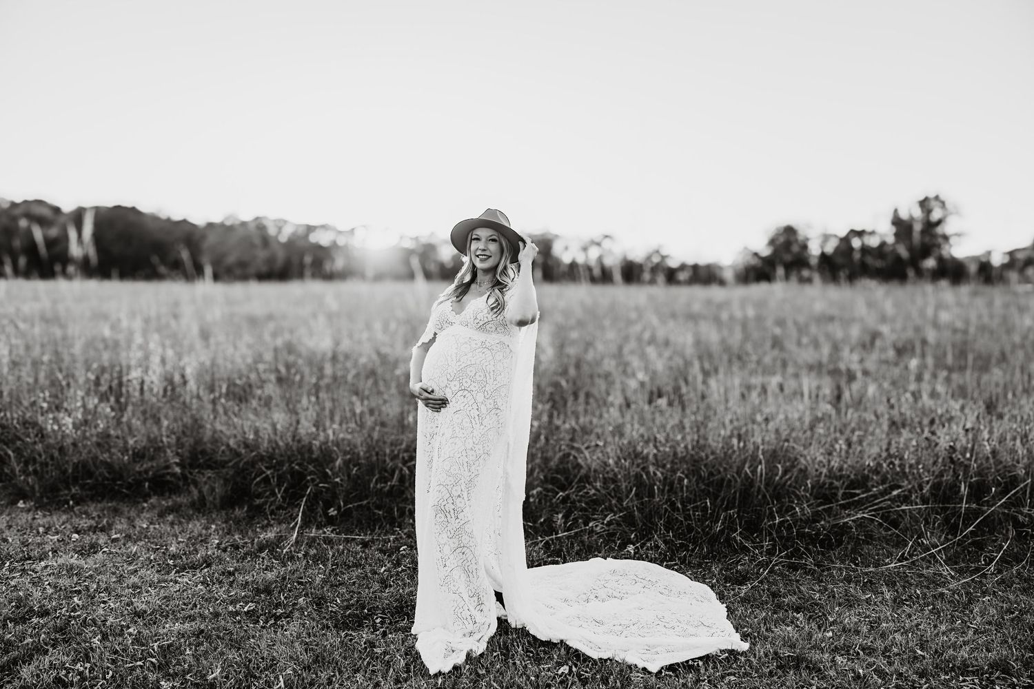 Black and white photo of pregnant woman in lace gown and hat standing in open field.