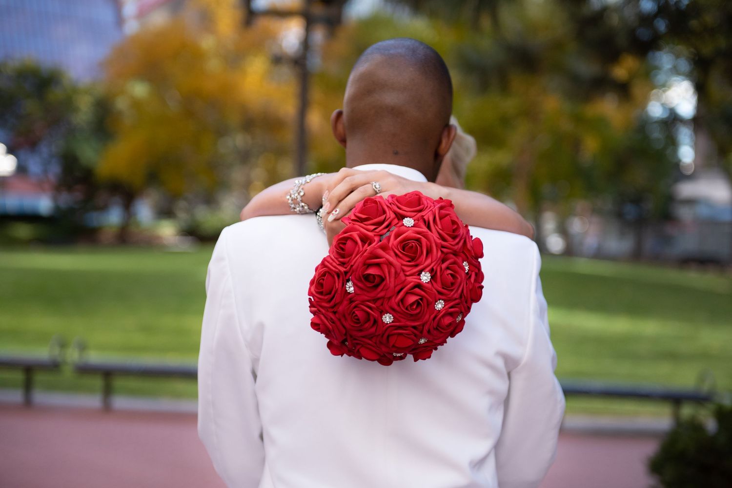 Bride in beaded gown placing hand on groom's shoulder while holding red rose bouquet from behind.