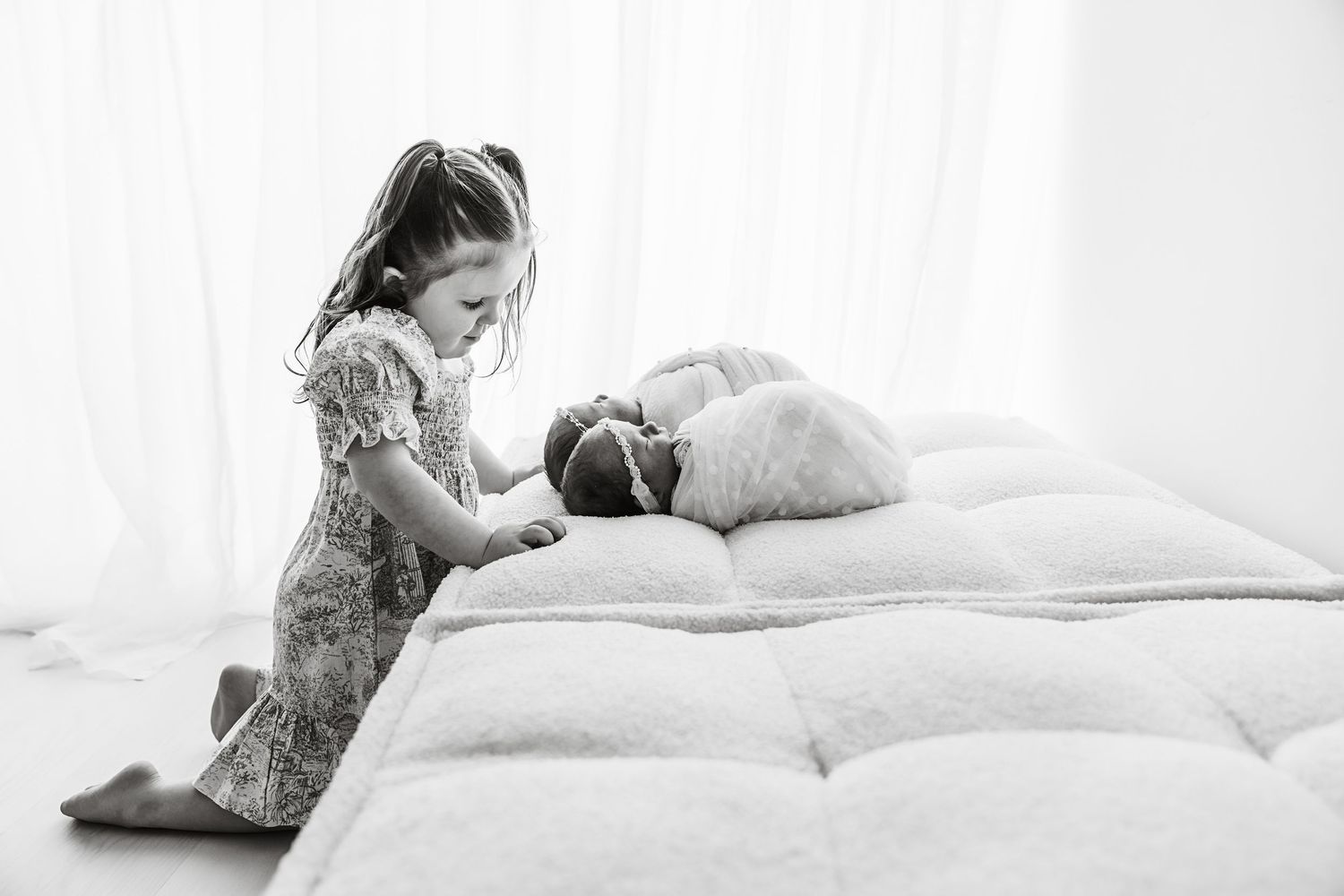 Black and white image of a little girl lovingly looking at a swaddled newborn on a mattress.
