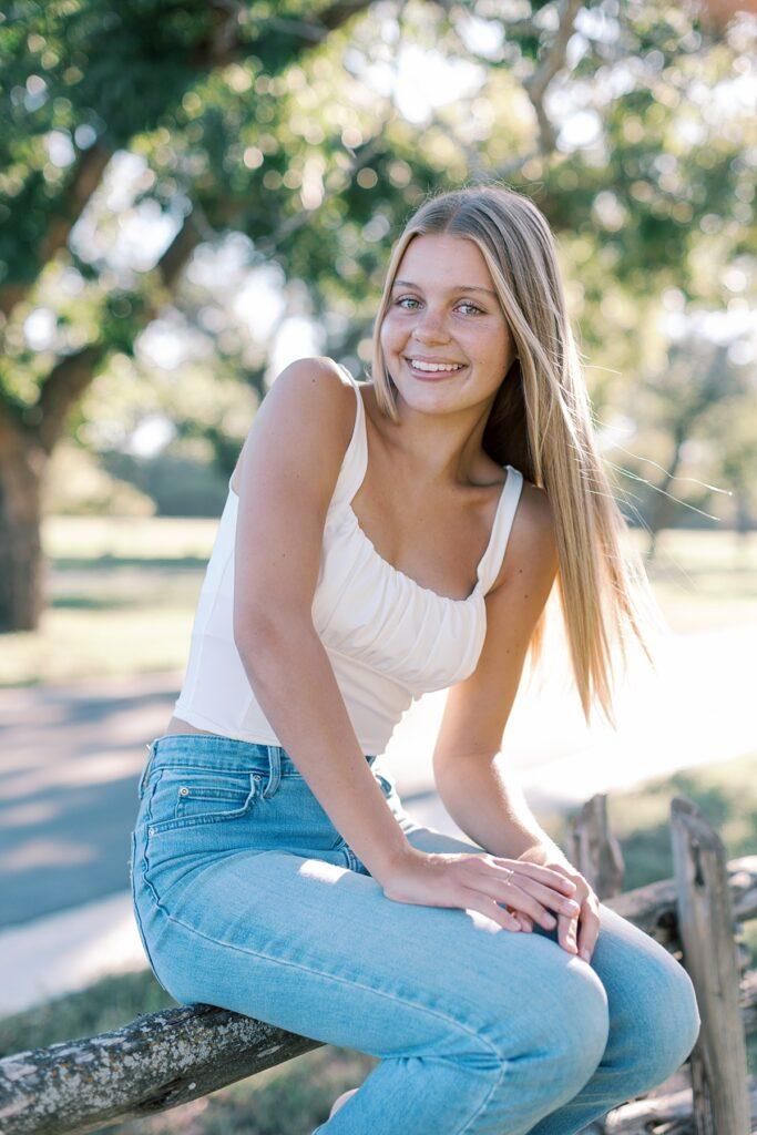 Smiling young woman with long blonde hair sitting on a wooden fence in jeans and white tank top outdoors.