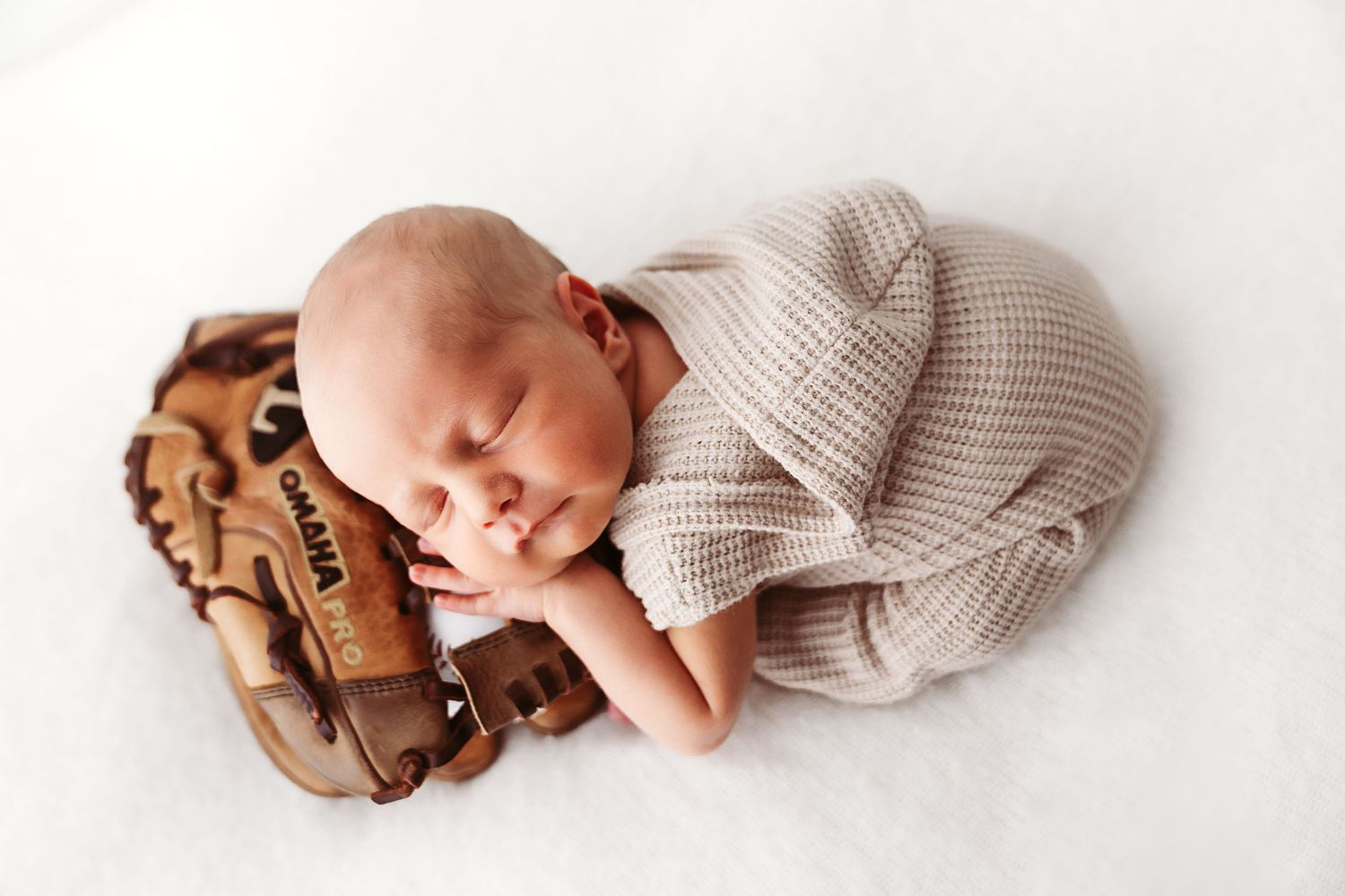 Sleeping newborn in beige knit outfit nestled in a vintage baseball glove.
