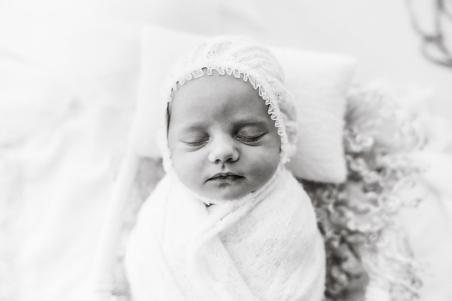 A black and white photo of a swaddled newborn baby wearing a lace bonnet sleeping on a soft pillow.