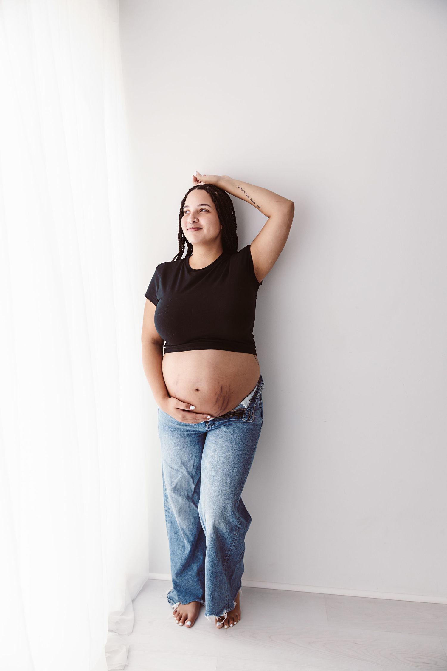 Pregnant woman in black crop top and jeans leaning against white wall with hand raised above head.