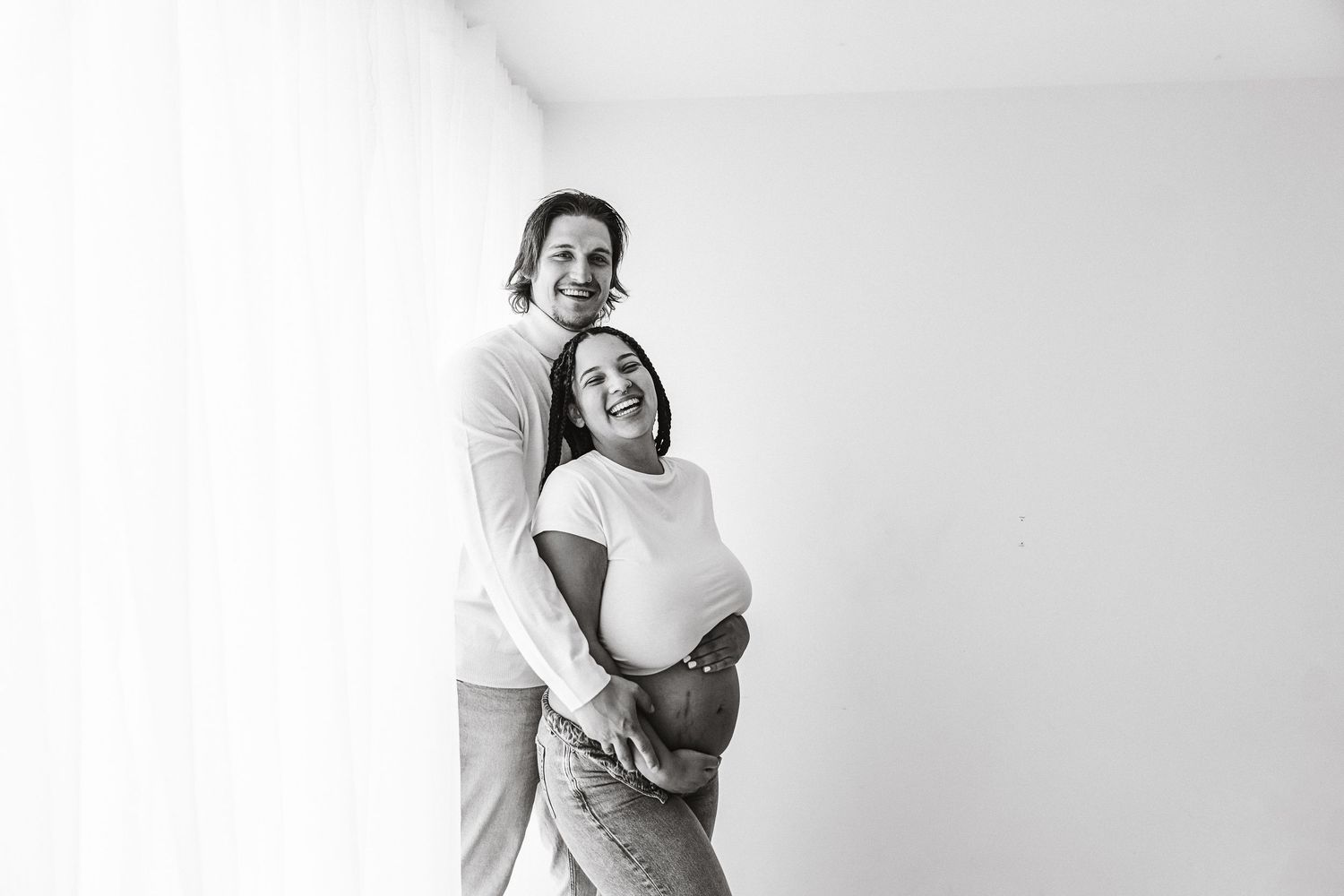 Black and white photo of smiling couple embracing pregnant belly by sheer curtain window.