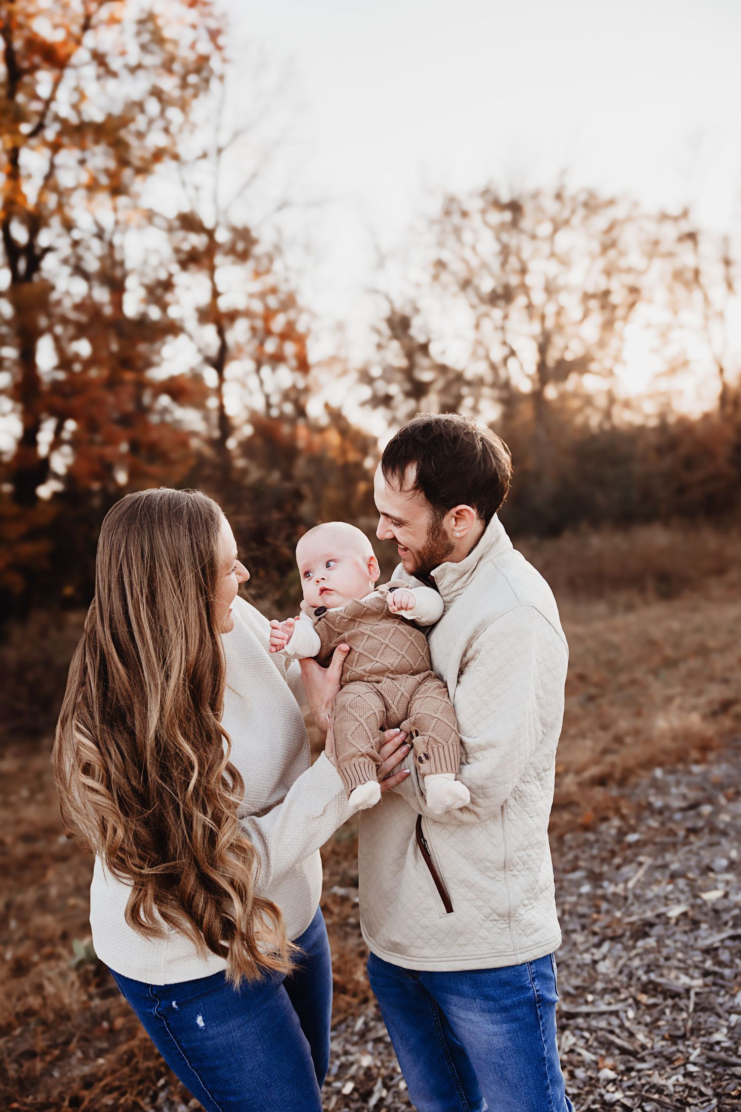 Family of three in autumn field, parents in cream sweaters holding their baby at golden hour.