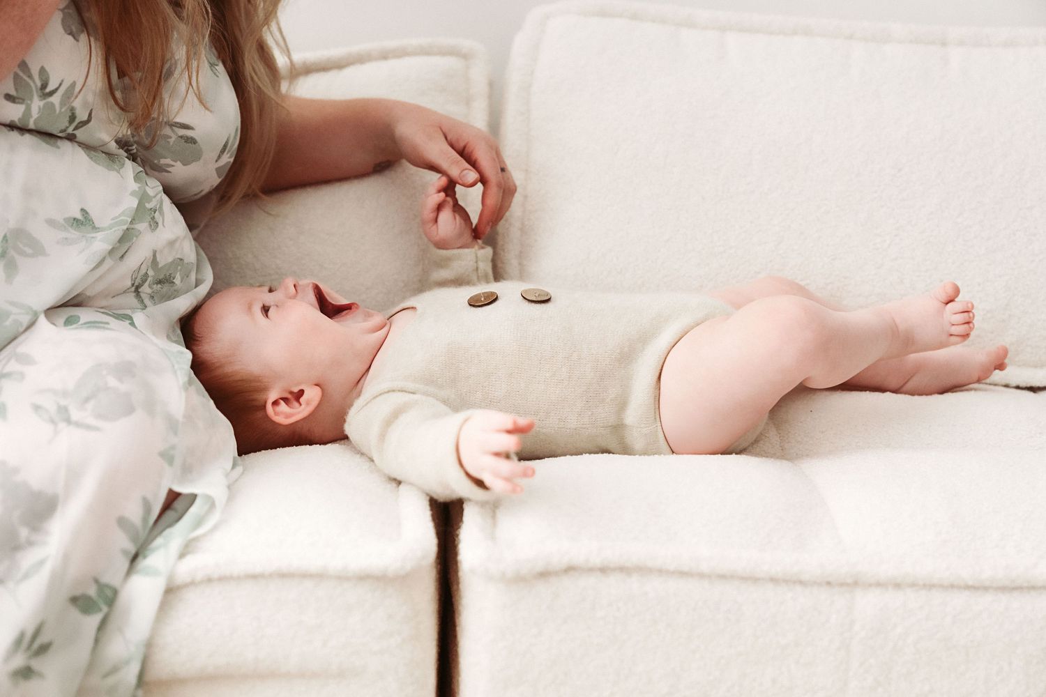 A mother sits beside her yawning newborn baby lying on a cream sofa.