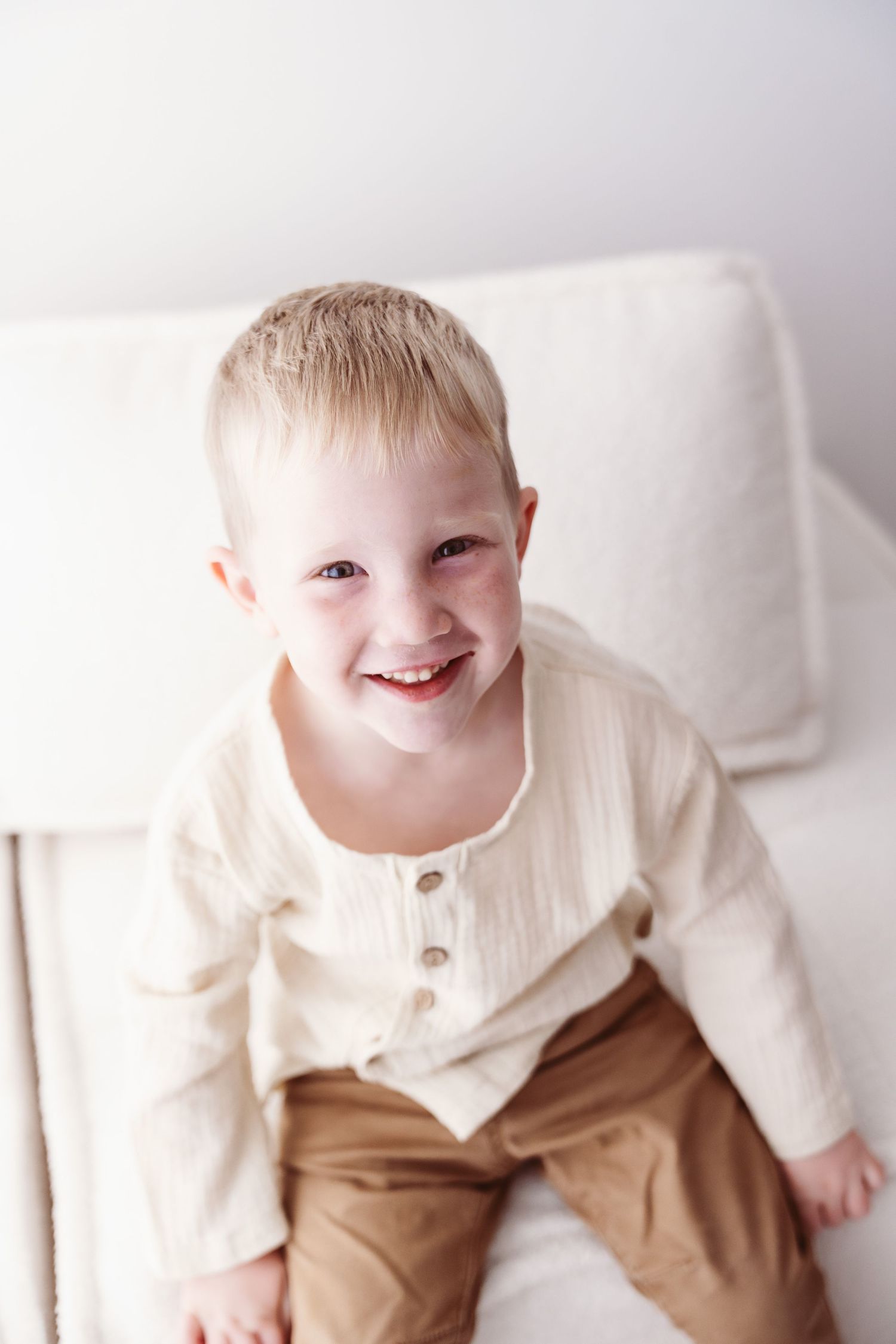 Happy smiling toddler boy in cream shirt and tan pants sitting on white couch.