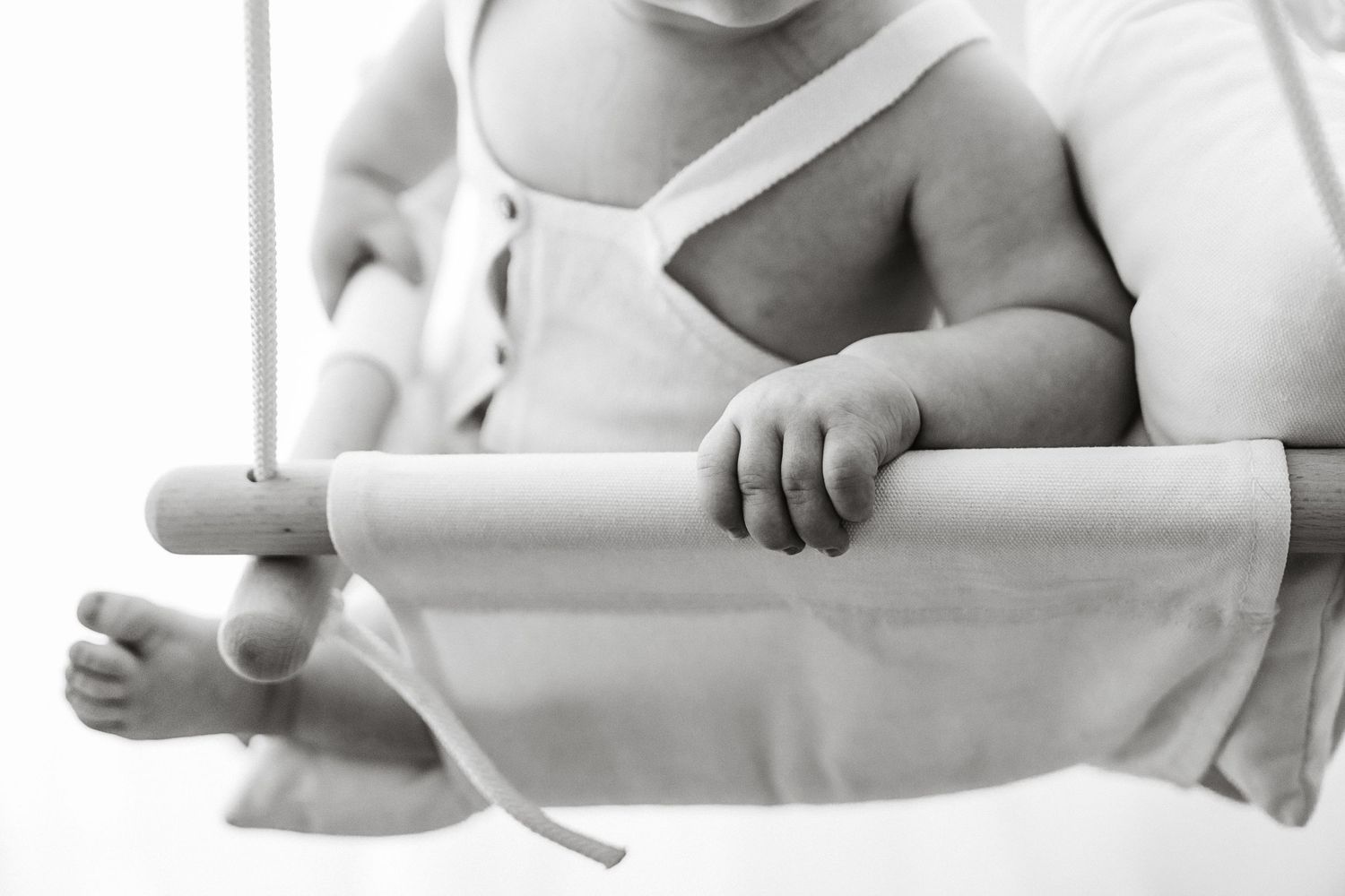 A close-up black and white photo of a baby gripping a wooden bar of a swing.
