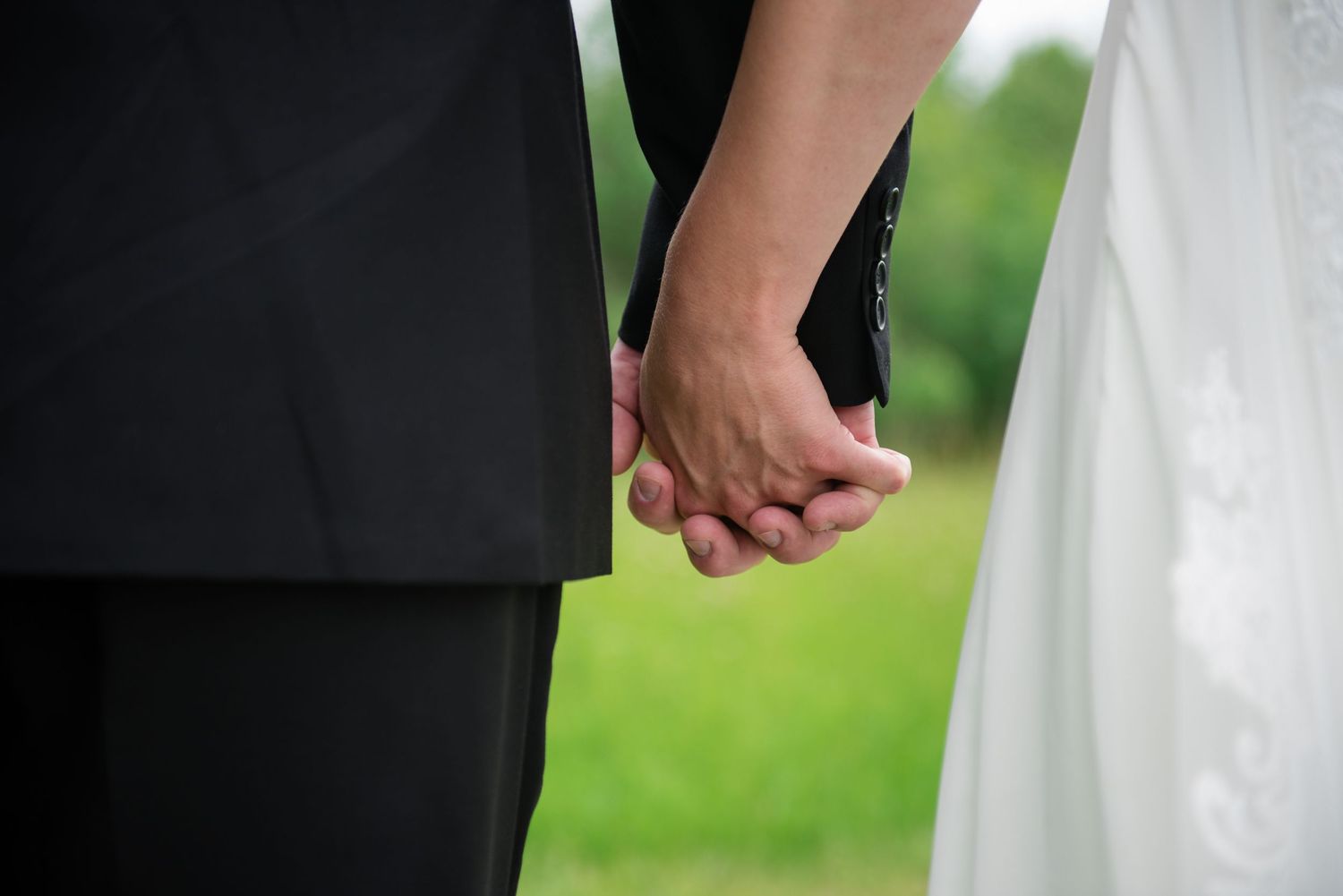 Bride and groom holding hands outdoors, dressed in black suit and white wedding gown against green backdrop.