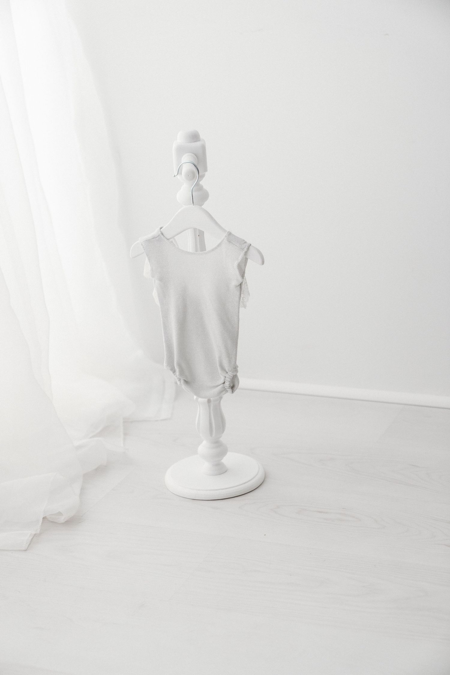 Minimalist gray sleeveless baby bodysuit displayed on a white decorative mini clothes stand in a bright white room.