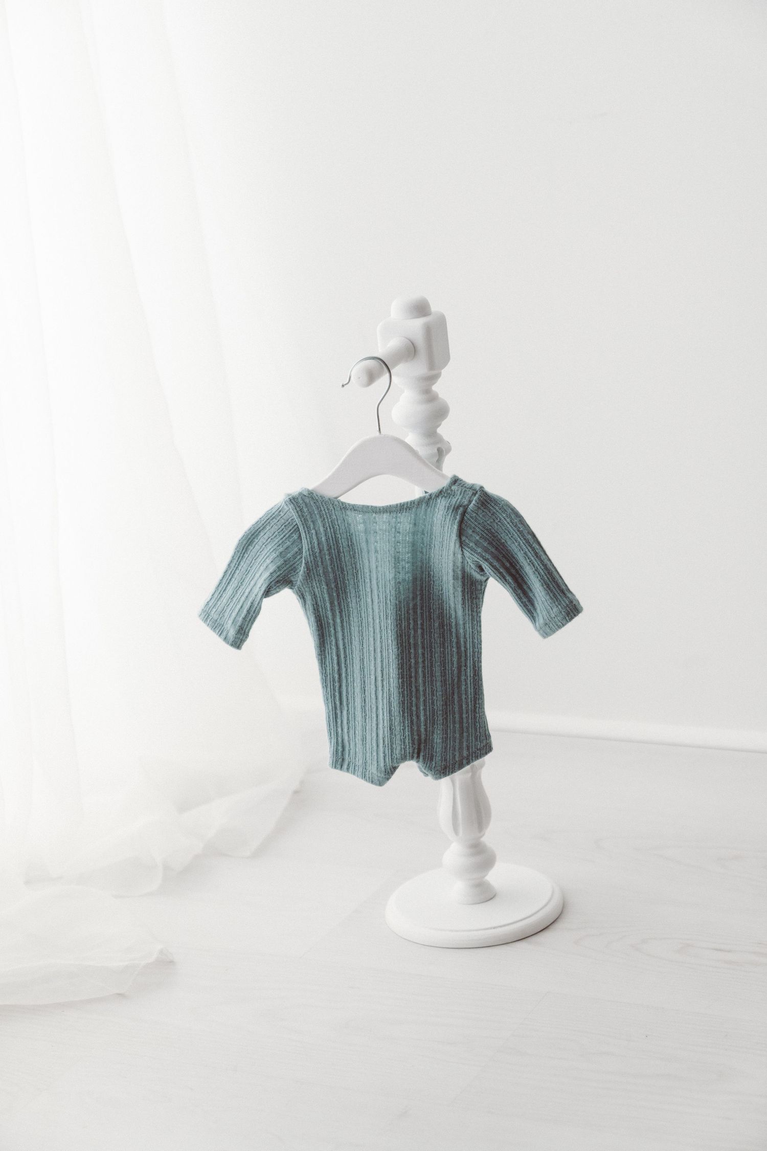 Teal ribbed baby bodysuit with short sleeves hanging on a white decorative stand against a white backdrop.