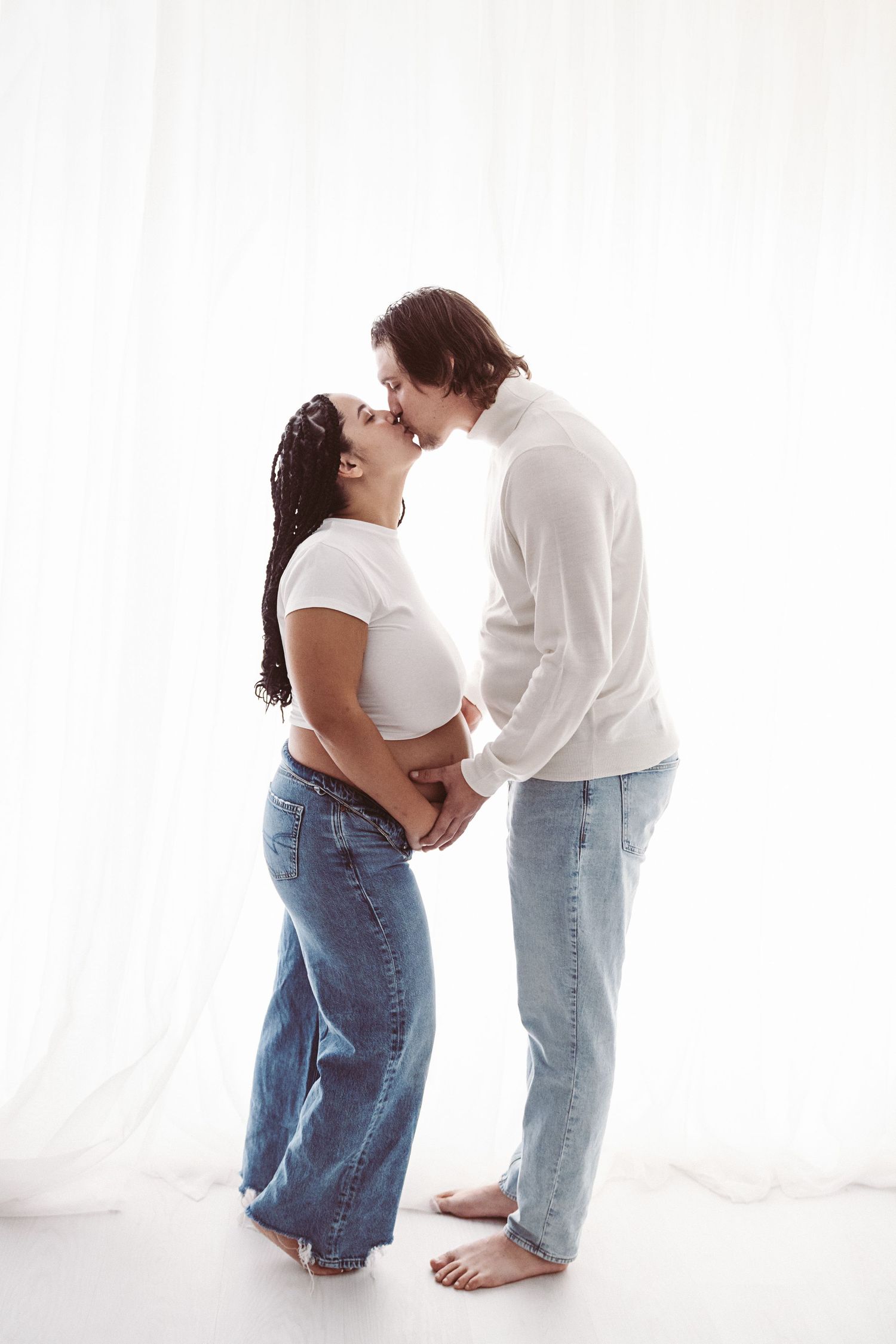 Pregnant couple sharing a kiss while holding baby bump together in white outfits against curtains.