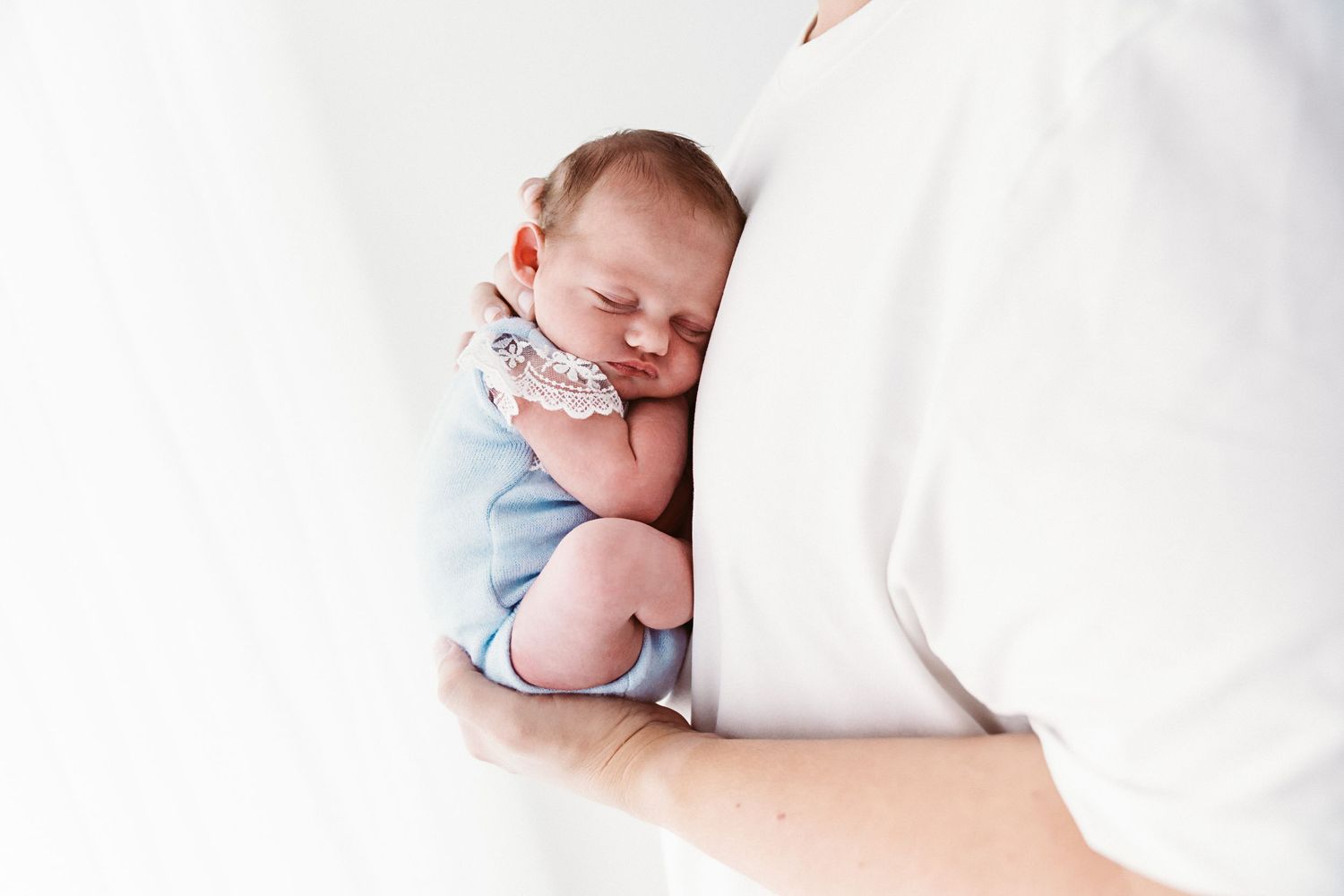 A newborn baby in a blue outfit sleeps peacefully against a parent's white shirt.
