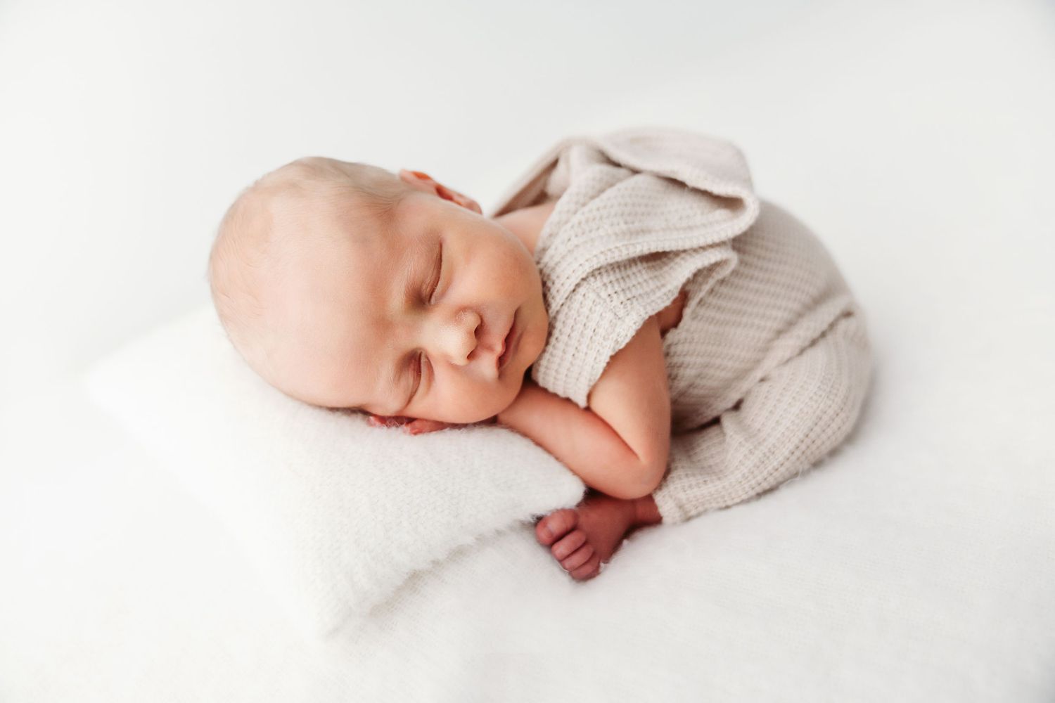 Sleeping newborn baby in a beige knit romper resting on a white pillow on white backdrop.