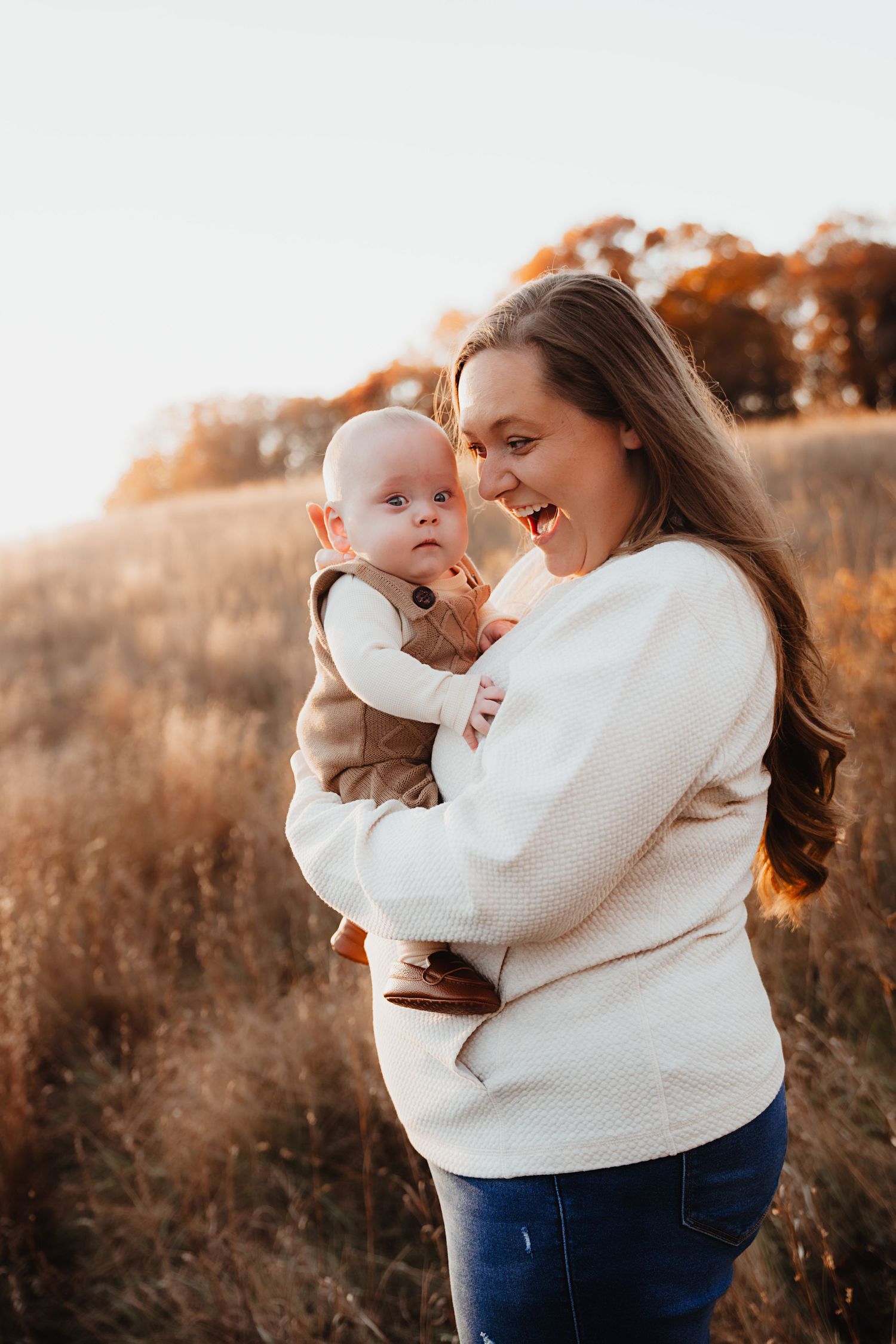 A smiling mother holds her baby in a sunlit autumn field, both wearing neutral tones.