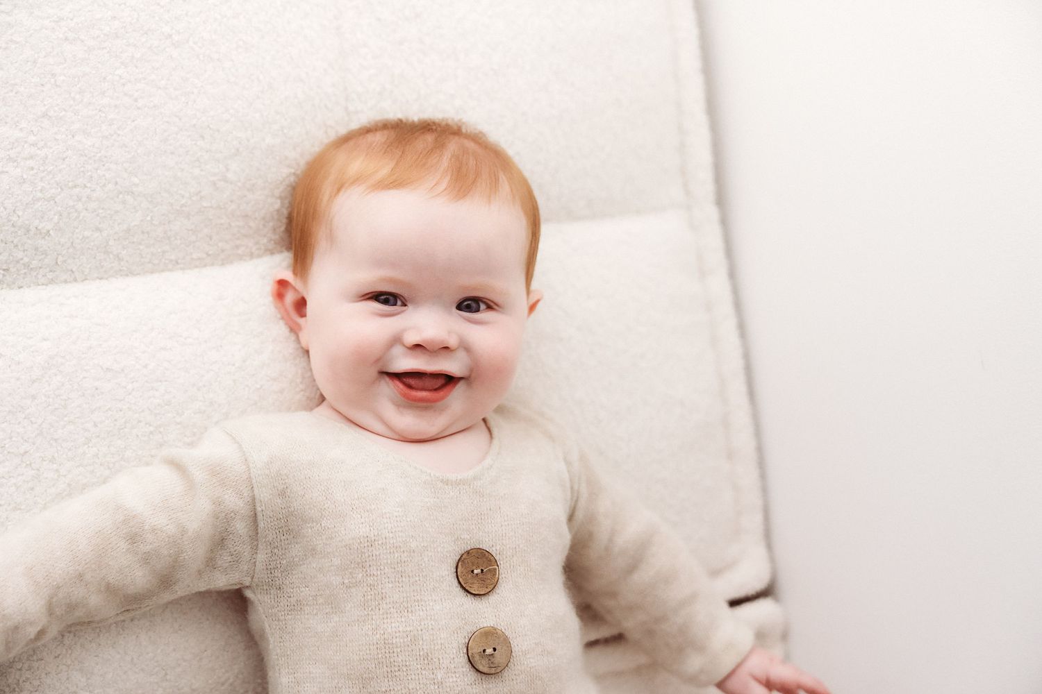 A smiling red-haired baby in a beige button cardigan lies on a white cushioned surface.