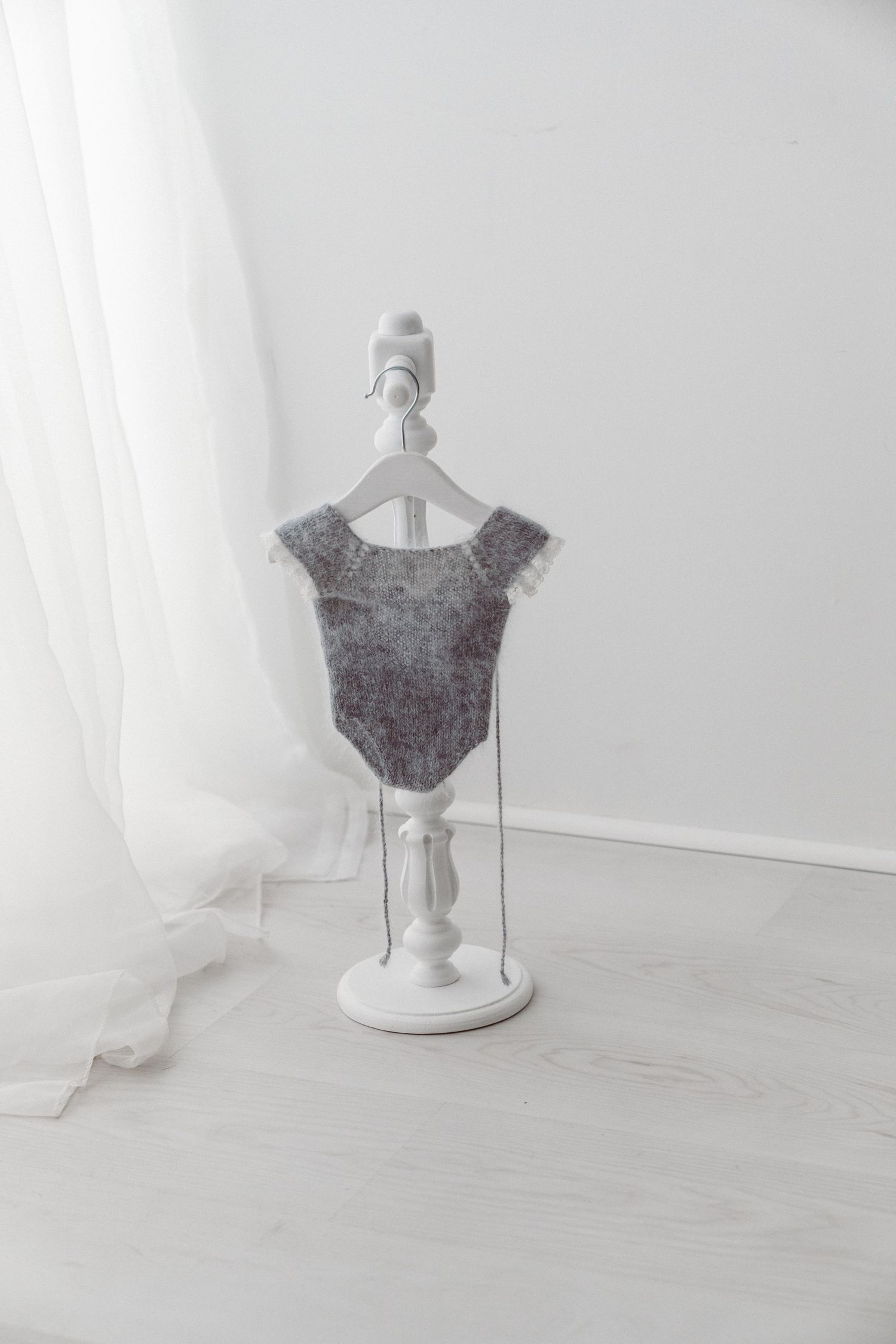Gray knitted baby bodysuit with delicate trim displayed on a white decorative mini clothes stand in a bright white room.