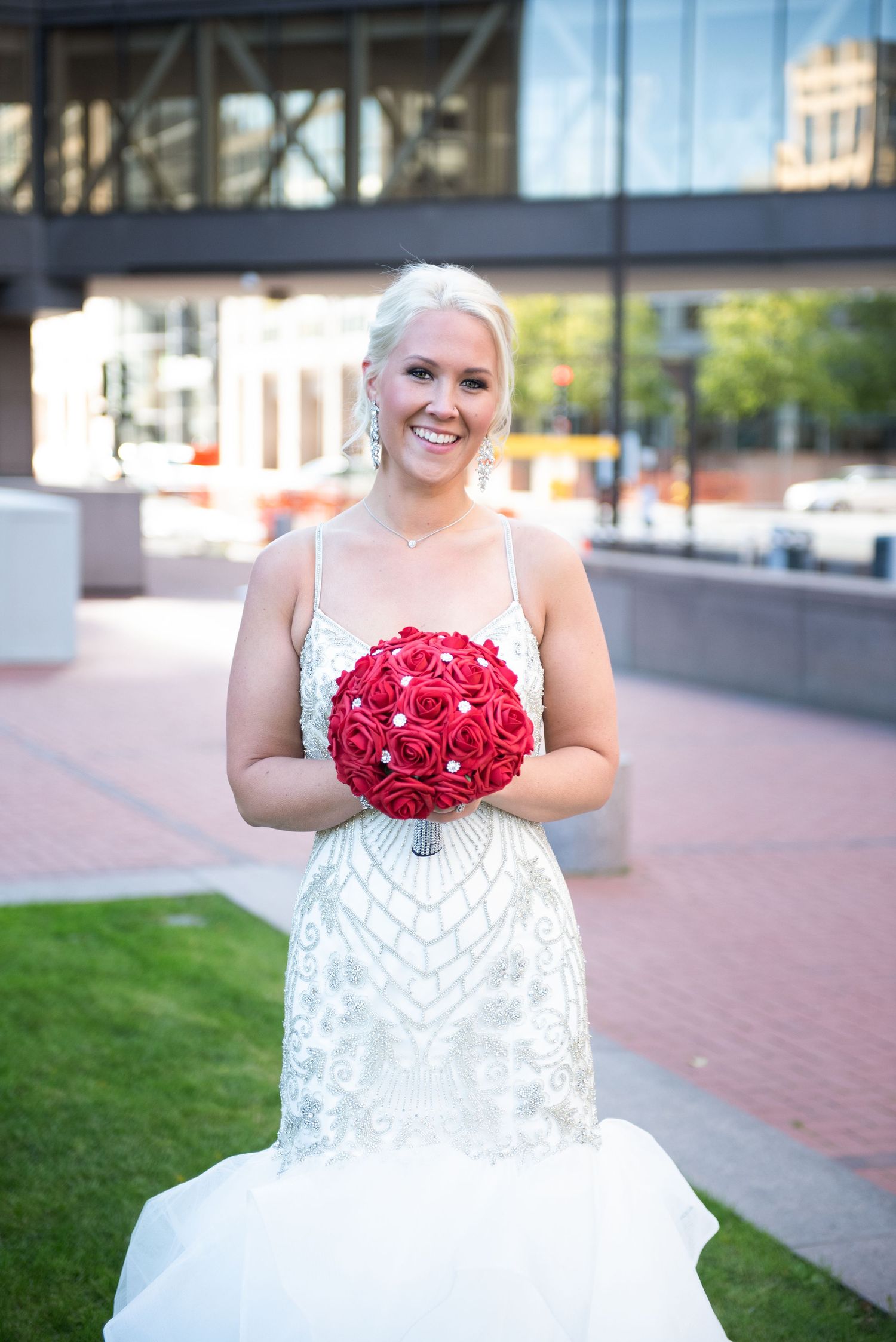 Smiling blonde bride in beaded gown holding a round red rose bouquet outside urban building.