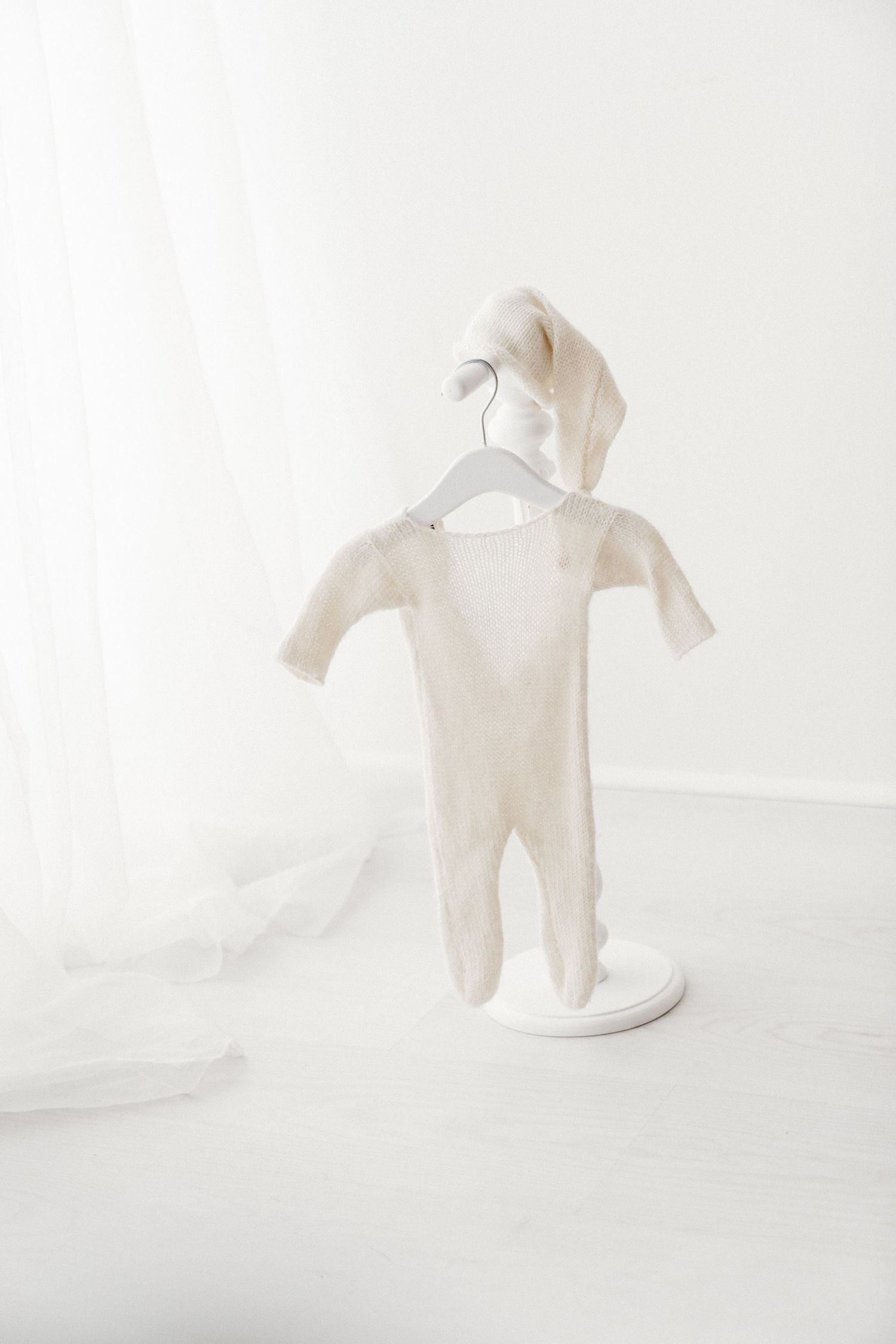 Cream knit baby footie pajamas with matching pointed hat displayed on a white decorative hanger stand.