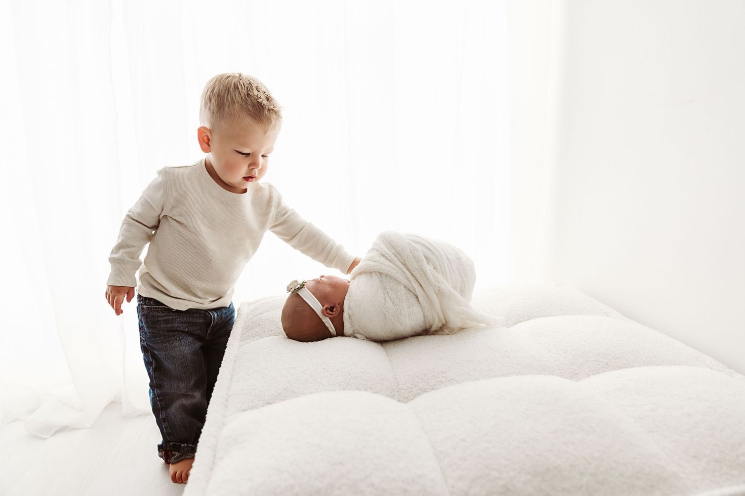 Curious toddler boy in white sweater gently touching swaddled newborn sibling on a white fluffy bed.