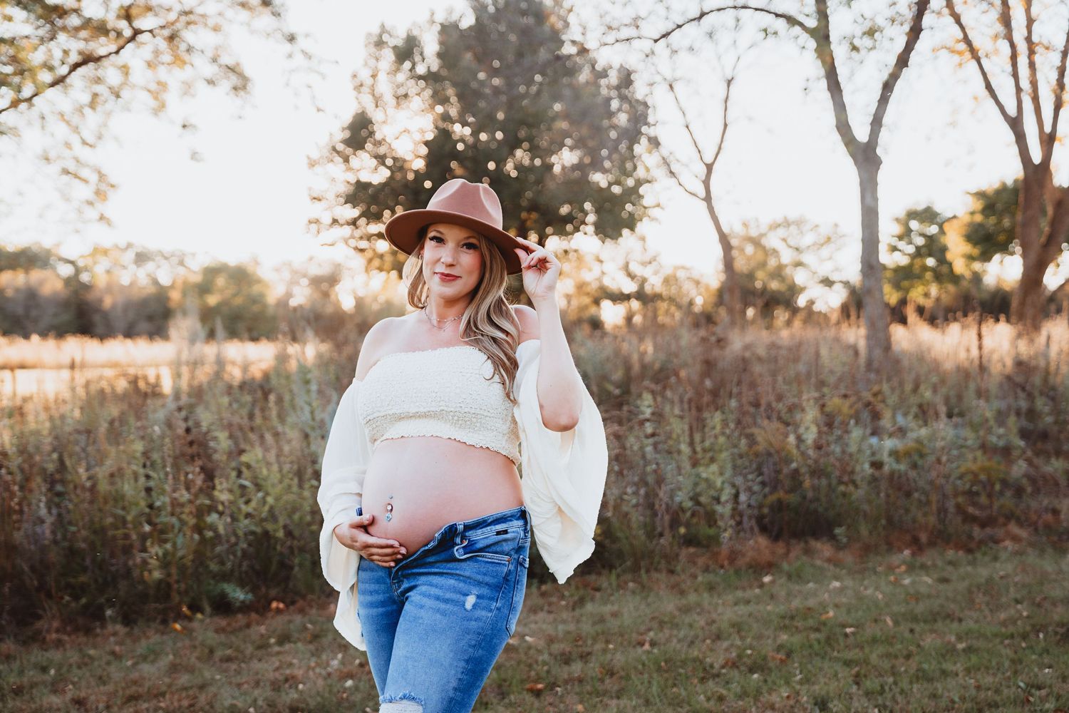 Glowing pregnant woman in a white off-shoulder top and brown hat holding her baby bump in an autumn field.