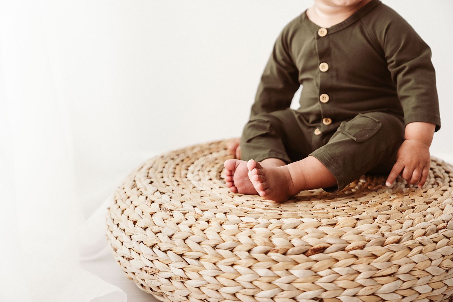 Close-up of baby in olive green romper sitting on a round woven rattan pouf.